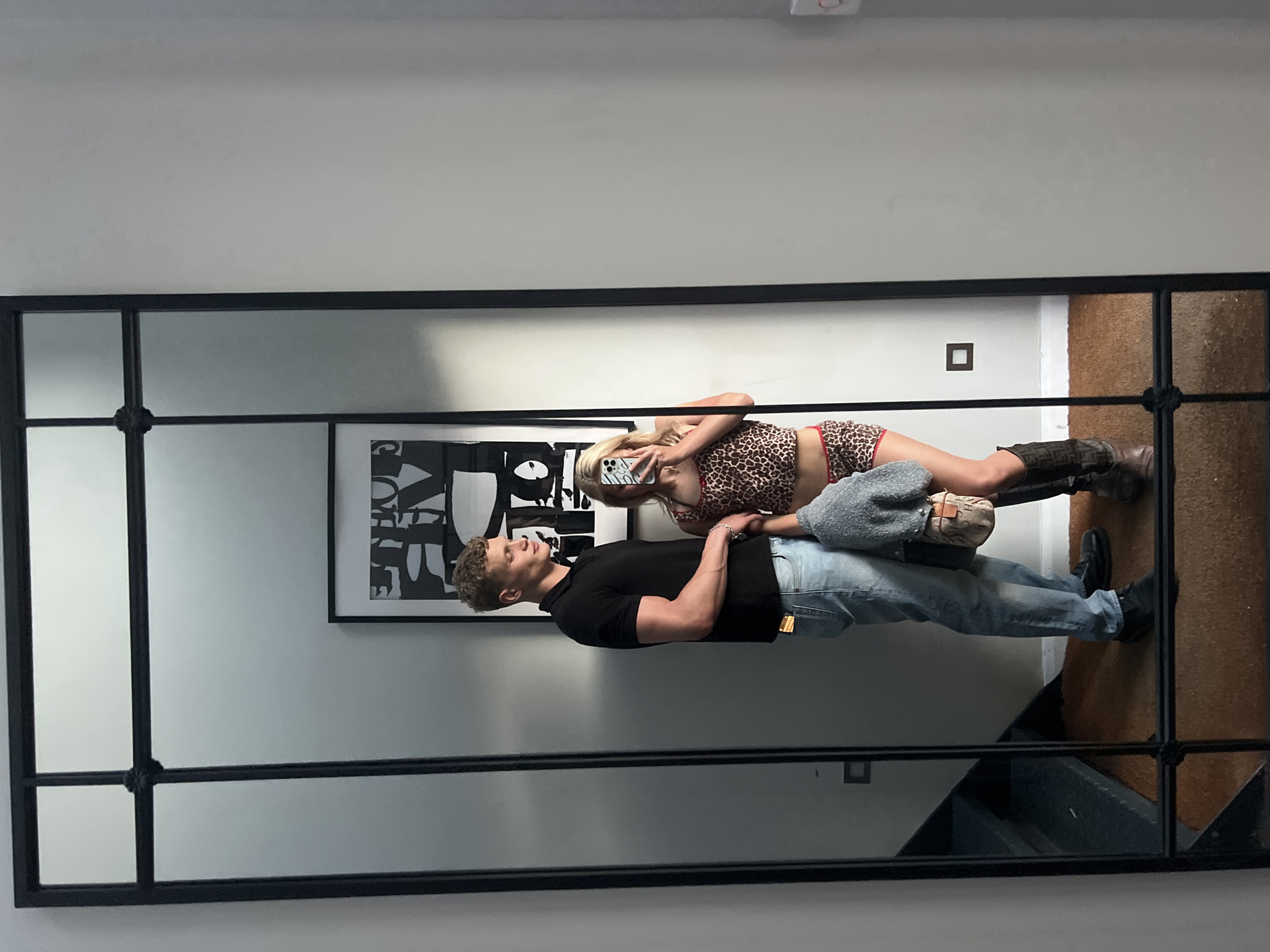 The image shows a reflection in a large, black-framed mirror. Reflected in the mirror are two individuals: a man and a woman. The man is standing, dressed in a black polo shirt and light blue jeans. The woman is reclining, wearing a cheetah print top and shorts, along with high boots. She is holding a cell phone in her hand. There is a piece of abstract art visible in the background. The reflection also reveals a staircase to the side. The location appears to be inside a building. There is no specific information to determine that the location is in the city of London, United Kingdom.