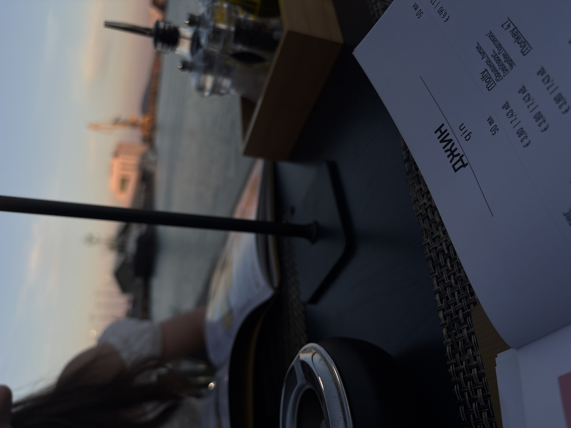 In Burgas, Bulgaria, a dimly lit outdoor dining scene unfolds at sunset. A person, partially visible and out of focus, is seated at a table. The table is set with an ashtray and a menu. In the background, a body of water, likely the sea, is visible, with a distant cityscape illuminated by the setting sun. The atmosphere is relaxed, with the focus on the table setting and the menu, hinting at a leisurely meal or drinks by the waterfront.