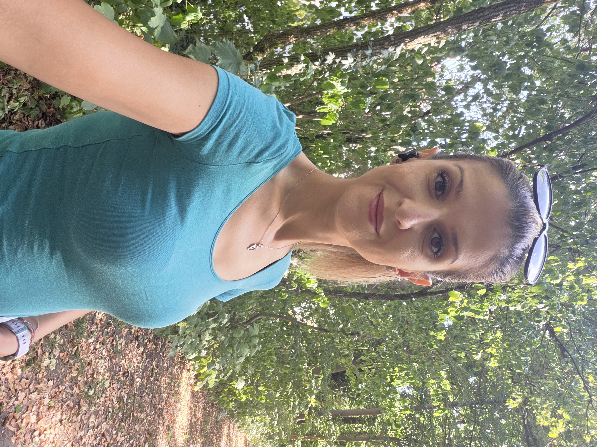 A woman is taking a selfie outdoors in a wooded area. She has blonde hair pulled back and is wearing a turquoise t-shirt, a silver necklace, and wireless earbuds. Sunglasses are perched on top of her head. The background consists of trees and a path covered in fallen leaves. The lighting suggests it is daytime. The exact location in Munich, Germany cannot be determined from the image.
