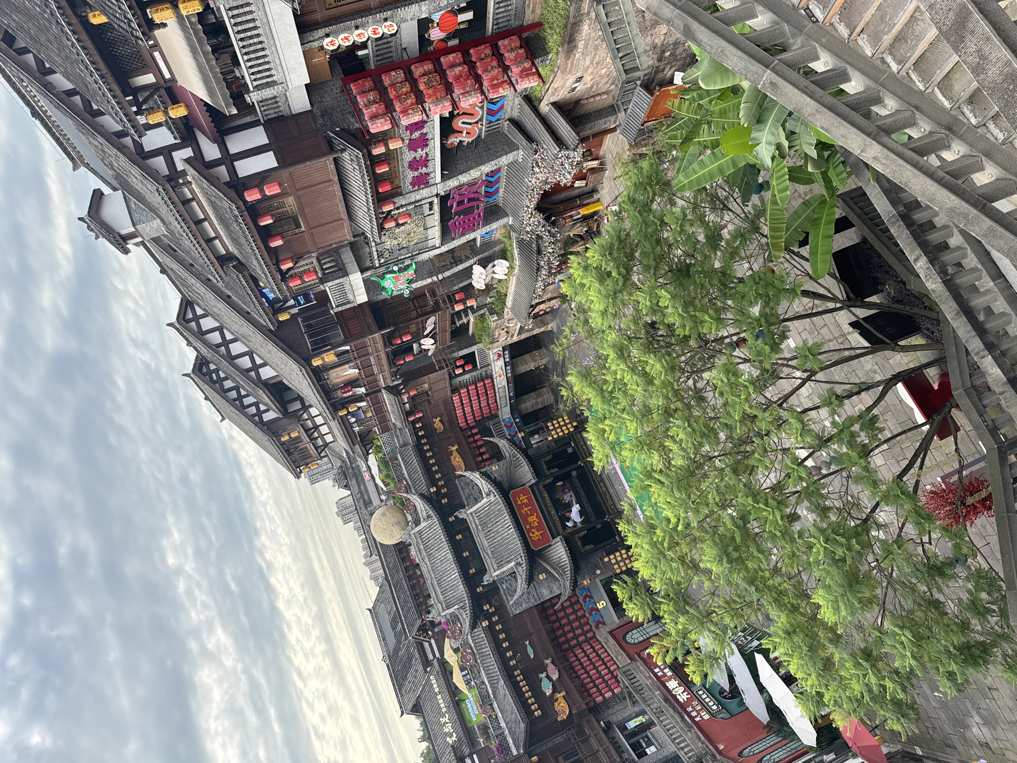 The image captures an aerial view of an urban scene in Shapingba District, China. The foreground features a lush tree with dense green foliage partially obscuring the view. To the left and below the tree, there is a gray concrete structure with a railing.
The architecture in the area is distinctively Chinese, characterized by traditional-style buildings with tiered, dark gray roofs. These buildings are adorned with red lanterns and various decorative elements. Some buildings have large signage in Chinese characters.
In the distance, a cloudy sky is visible, suggesting either morning or late afternoon lighting. The scene appears to be a commercial or tourist area with numerous shops and restaurants. There are people visible in some areas, suggesting activity and pedestrian traffic.