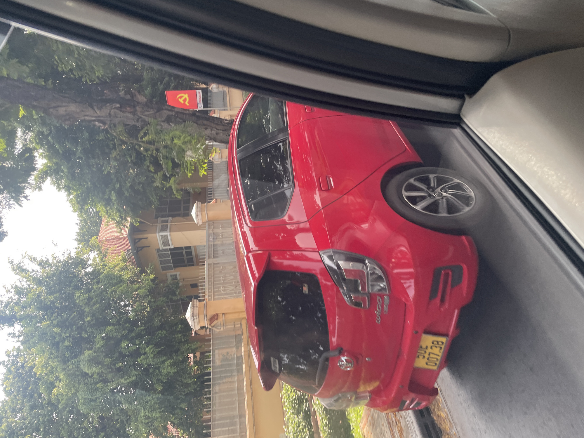 A bright red Toyota Wigo is parked on a street, its rear bumper facing the camera. The car's license plate, a yellow rectangle with black lettering, reads "30E 007.38". The car is situated in front of a beige building with a white fence. A tall tree with dense green foliage stands to the right of the car, and a red flag with a yellow hammer and sickle symbol is visible behind the tree. The scene appears to be in Thủ Đức City, Vietnam, as indicated by the license plate format.