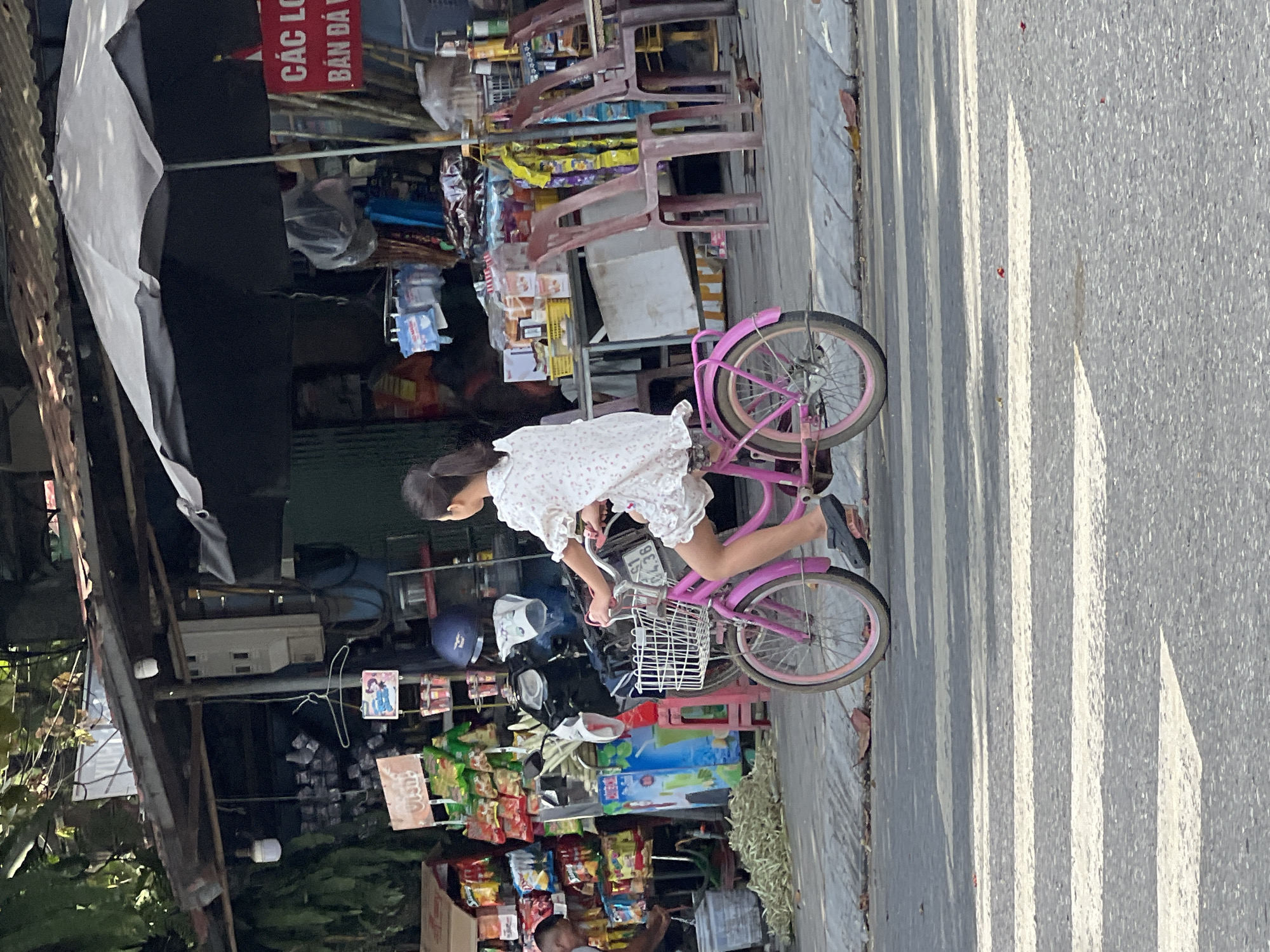 A young girl with dark hair pulled back in a ponytail, wearing a white floral dress, rides a pink bicycle with a white basket on a street in Tân Quang Commune, Vietnam. The girl is facing away from the viewer and to the left, her feet on the pedals. The bicycle has pink rims. The background shows a bustling street scene with a covered market stall selling various goods, including snacks and drinks. A motorcycle is parked behind the stall. Several plastic chairs are visible in front of the stall. A red sign with white text, appearing to be Vietnamese, is partially visible to the right. The street has a pedestrian crossing marked with white stripes. The lighting suggests it is daytime, with some shadows cast on the road.