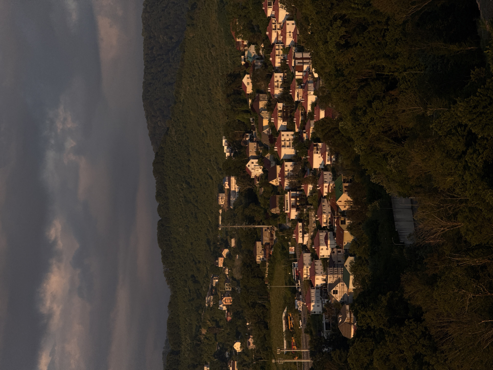 The image captures a view of a residential area on a hillside in Sochi, Russia, bathed in the warm, low light of either sunrise or sunset. The scene is dominated by a dense, verdant forest in the foreground and midground, with trees of varying heights and foliage density.
Scattered amongst the trees and along the slopes are numerous houses, many of which appear to be villas or detached homes. These residences are predominantly characterized by white or light-colored facades and red-tiled roofs, creating a distinct visual pattern across the hillside. Some of the houses are clustered together, while others are more isolated, suggesting a sprawling development.
In the background, a larger, darker, forested mountain range rises, its contours softened by the atmospheric conditions. Above the mountains, a dramatic sky is visible, filled with heavy, dark clouds interspersed with areas of lighter, possibly sunlit, cloud formations. The lighting casts long shadows and highlights, giving the scene a sense of depth and a slightly moody atmosphere.
A road or highway is visible in the midground, with several vehicles, including buses and cars, traveling along it. There are also some utility poles and lines, indicating infrastructure within the area. The overall impression is of a peaceful, somewhat rural residential community nestled within a natural, mountainous landscape.