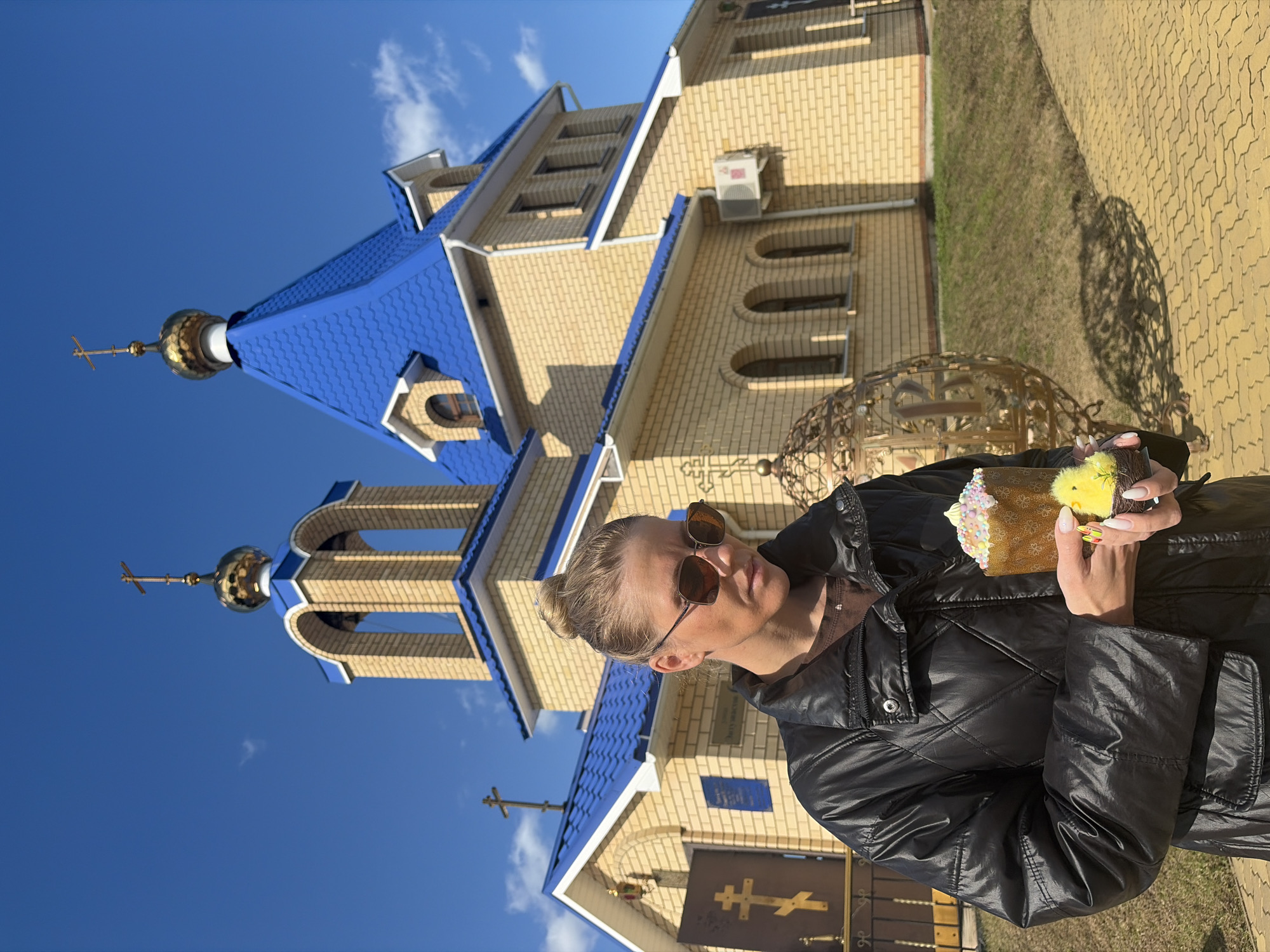 A woman stands in front of the Orthodox Church of St. Sergius of Radonezh in Chaikovsky, Russia, holding an Easter cake and a chick toy. The church features yellow brick walls and a blue roof, with crosses atop its domes. The sky is blue, and sunlight casts shadows on the brick sidewalk. The woman wears sunglasses and a black jacket.