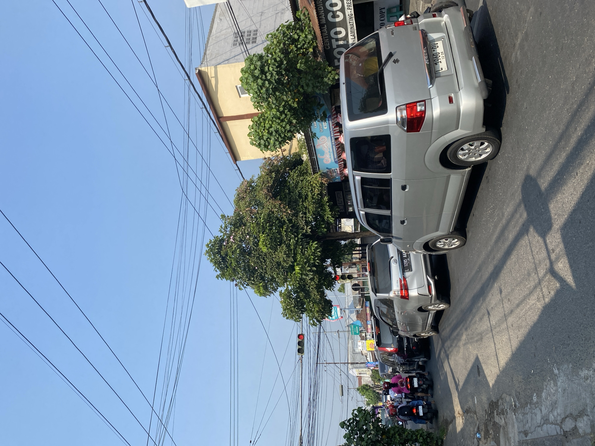 The image shows a traffic jam on a city street in Kediri City, Indonesia, on a clear day. Several cars and motorcycles are stopped on the road, suggesting congestion. Overhead power lines crisscross the sky, and traffic lights are visible, with at least one showing a red signal. Buildings line the street, including a yellow and gray multi-story building with businesses on the ground floor, featuring signage for "CENTRAL MANDIRI FOTO COPY" and "Sweet." Green trees are scattered along the roadside, providing shade. The perspective is from a slightly elevated position, looking down the street.