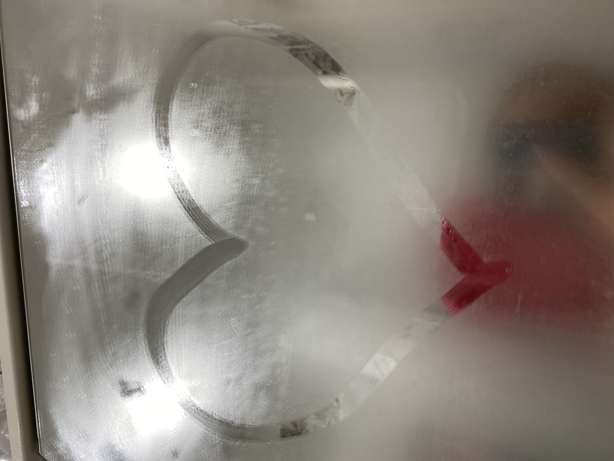 A hazy, indoor shot focuses on a fogged-up glass surface. A heart shape has been drawn into the condensation, with its outline appearing clearer against the frosted glass. A blurred figure in a red shirt is faintly visible through the glass, near the right side of the image. The background is indiscernible due to the fogging. The scene likely takes place in a bathroom due to the fog on the glass, but the exact location in Chaikovsky, Russia is not discernible from the image.