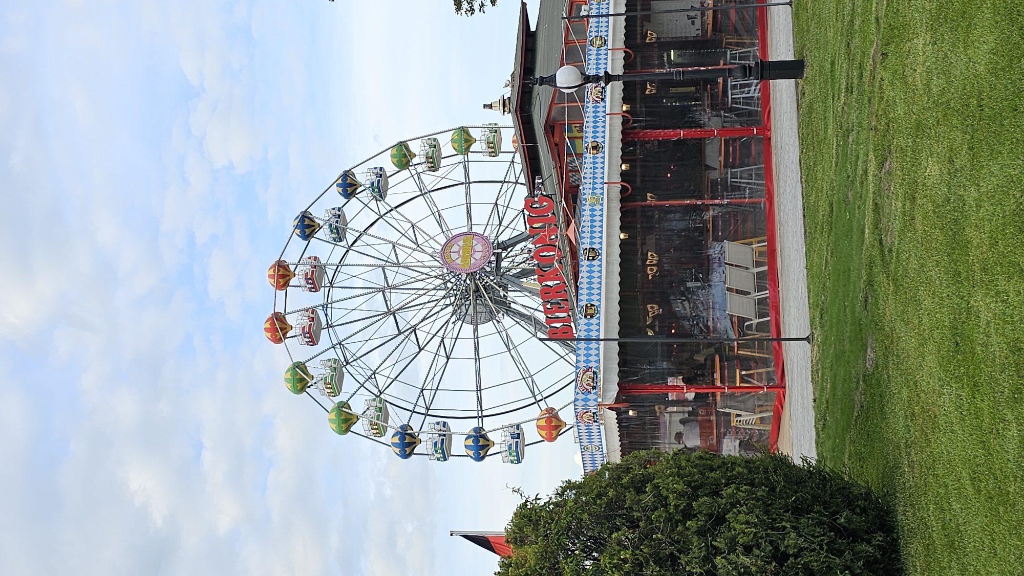 The image shows a Ferris wheel called "BERGKRONE" next to an adjacent building in the city of Varna, Bulgaria. The Ferris wheel has colorful gondolas. The adjacent building has a red frame and a reflective facade, showing the interior with tables and chairs. There is a patch of green grass and shrubbery in the foreground, and the sky in the background has scattered clouds. Lampposts stand between the Ferris wheel and the building.