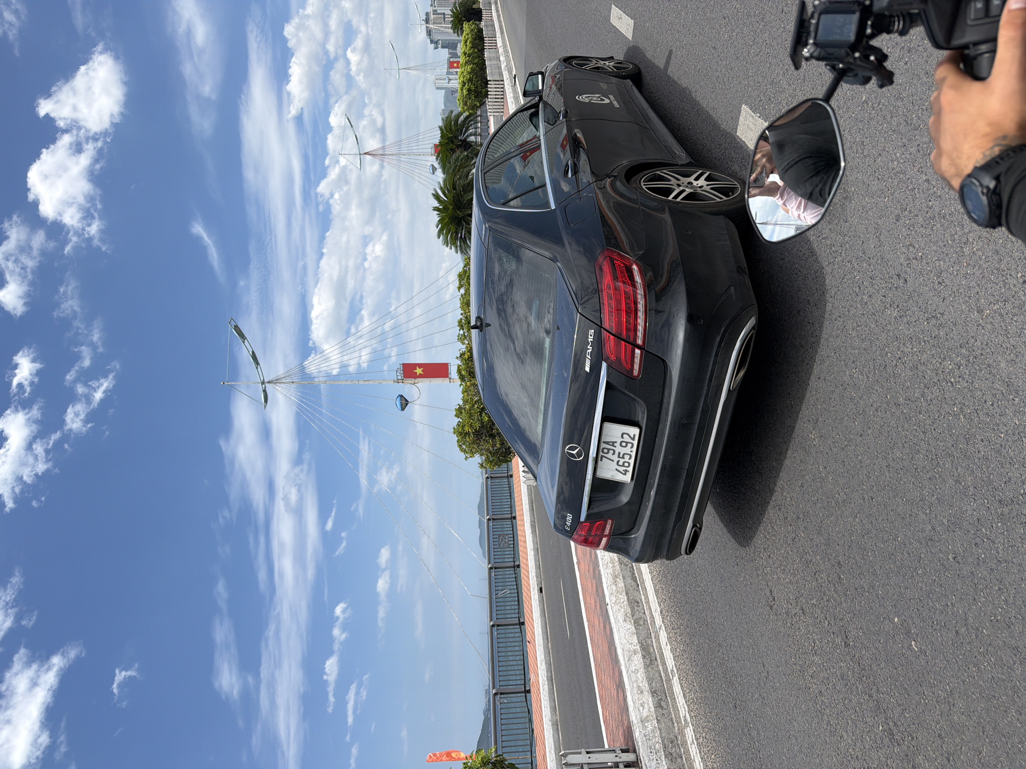 The image captures a street-level view of a black Mercedes-Benz E400 sedan driving on a road in Nha Trang, Vietnam. The car, with license plate 79A 465.92, is positioned on the right side of the road, and the perspective suggests it is being followed by a motorcycle, as a rider's hand and a motorcycle mirror are visible in the foreground.
The backdrop features a bridge with distinctive architectural elements, including tall, white masts with cables extending from them, and red triangular structures. A flag with a yellow star on a red background, the flag of Vietnam, is prominently displayed on a mast to the left. The bridge spans over water, with a railing and a pedestrian walkway along the side. Palm trees line the walkway, and a cityscape with buildings is visible in the distance. The sky is bright blue with scattered white clouds, indicating a sunny day. The road itself is asphalt, with white lane markings. A red and white striped concrete barrier is present on the left side of the road.