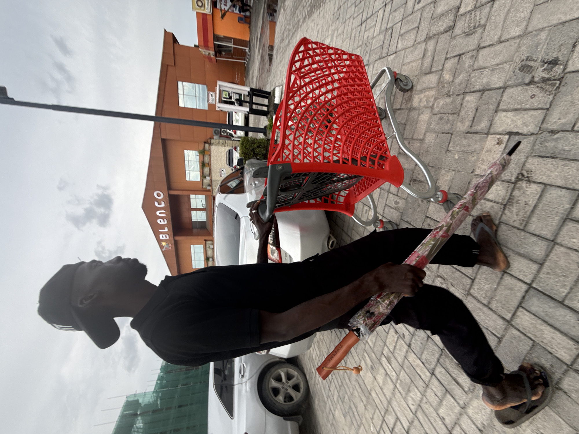 A Black man, wearing a black t-shirt, black pants, and a black baseball cap, walks past a red shopping cart in front of a building in Ado, Nigeria. The man is carrying a patterned umbrella in his right hand. The building behind him has an orange facade and large windows. There are cars parked in the background, and the ground is paved with cobblestones. The sky is overcast.
