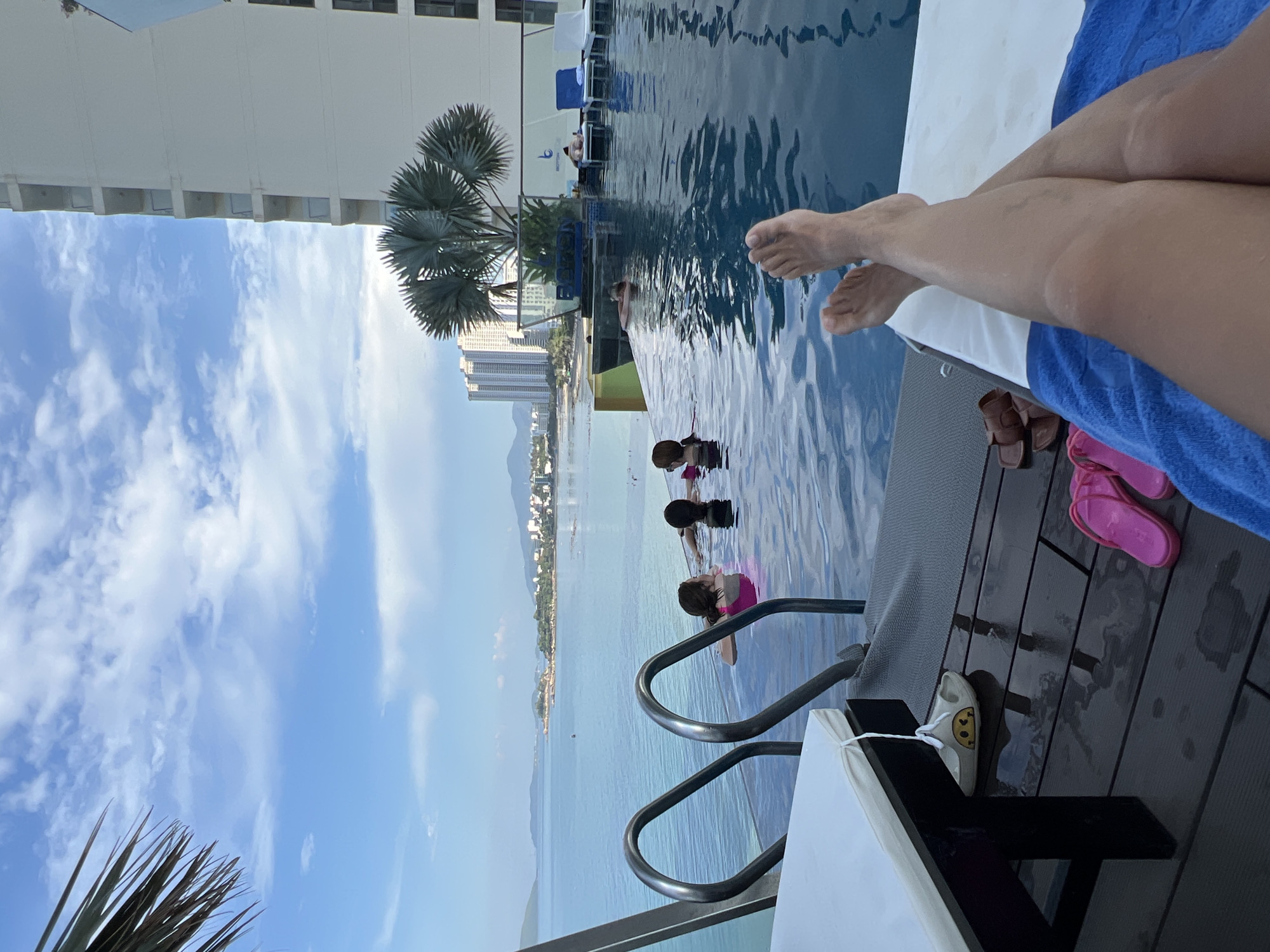 This is a photograph taken at an infinity pool overlooking the ocean, likely in Nha Trang, Vietnam, given the context. The viewpoint is from someone lounging by the pool, with their legs extended into the frame. The image is oriented vertically, with the sky and buildings appearing on the left and the pool and deck on the right.
The pool itself is a prominent feature, its blue water reflecting the sky. Several people are visible in the pool, including at least two children and an adult. They appear to be swimming or relaxing in the water. In the background, a palm tree stands tall, with a modern hotel or apartment building behind it. Further in the distance, a city skyline with tall buildings and a mountainous landscape is visible, suggesting an urban coastal setting.
On the wooden deck beside the pool, there are a few objects: a pair of brown sandals, a pair of pink flip-flops, and a white sandal with a smiley face design. A blue towel is also visible, draped over a lounge chair, and the legs of the person taking the photo.
The sky is a bright blue with scattered white clouds, indicating a sunny day. The water in the pool is calm, with gentle ripples. The overall mood of the image is relaxed and vacation-like. There are no visible text elements in the photograph.