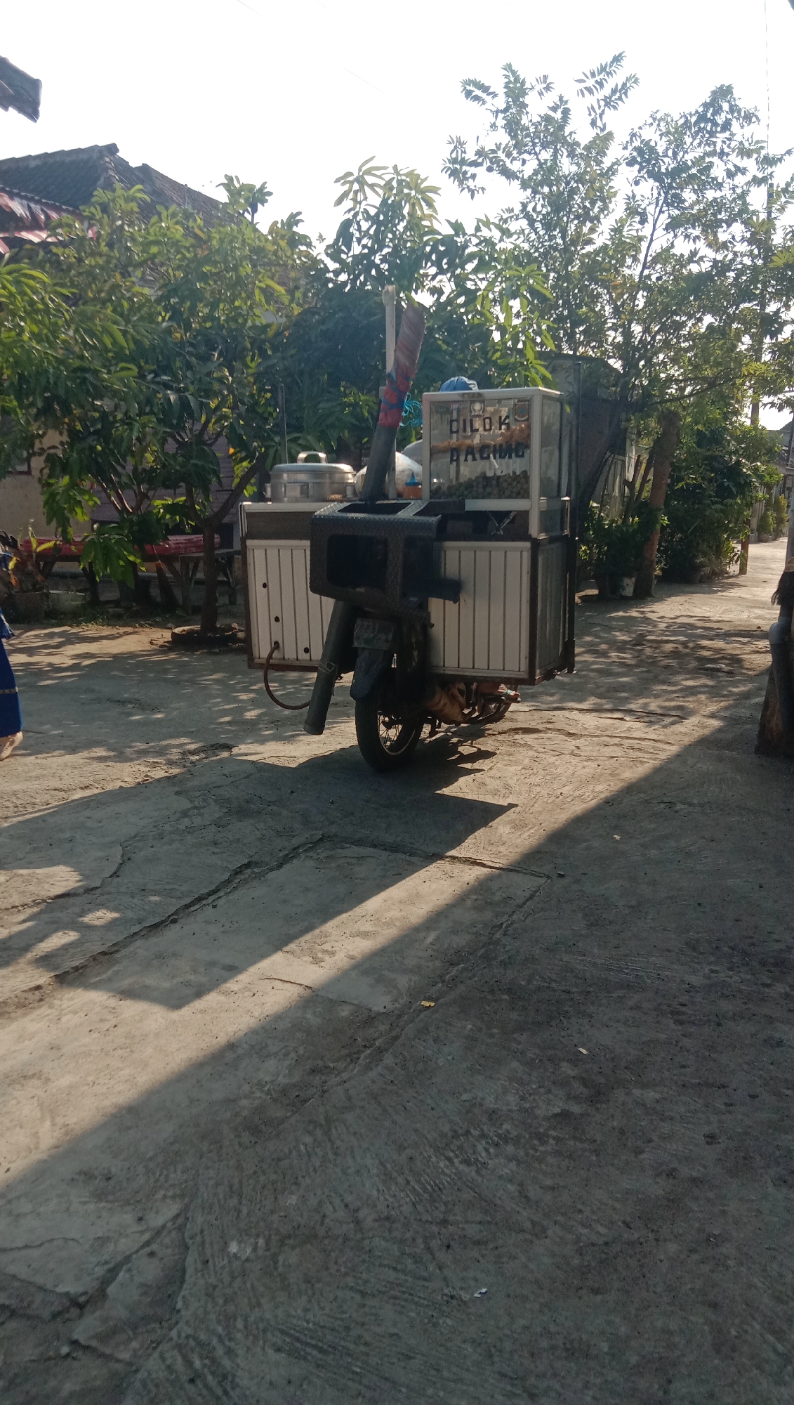 The image shows a motorcycle with a food cart attached to the back, parked on a concrete pathway in the city of Sidogiri, Indonesia. The food cart appears to be selling meatballs, indicated by the text "DILOK BAKSO" visible on its side. The motorcycle is a dark color and has a metallic grill structure mounted on top of the food cart, possibly for cooking or display. The surrounding environment includes greenery, trees, and residential buildings, suggesting a semi-urban or rural setting. The sun is casting long shadows, indicating it is either early morning or late afternoon. No people are actively engaged with the food cart or motorcycle.