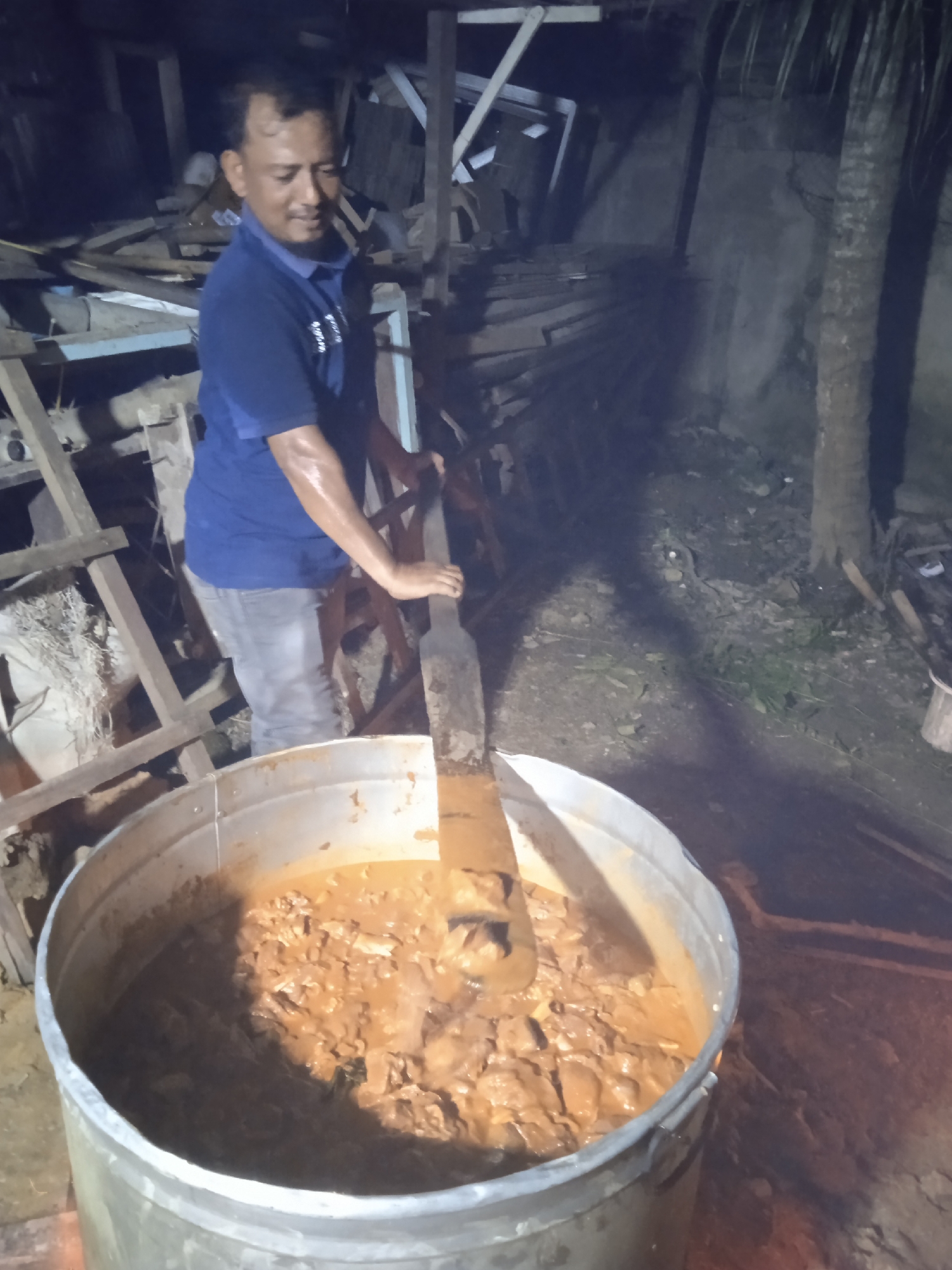 In Keudeteupinpunti, Indonesia, a man in a blue t-shirt and jeans stands over a large metal pot, stirring its contents with a wooden paddle. The pot is filled with a brown, stew-like mixture containing visible pieces of meat and vegetables. The scene is set outdoors at night, with a dimly lit background showing construction debris, wooden structures, and a palm tree. The ground is uneven and appears to be dirt. The man's face shows a slight smile as he concentrates on the task.