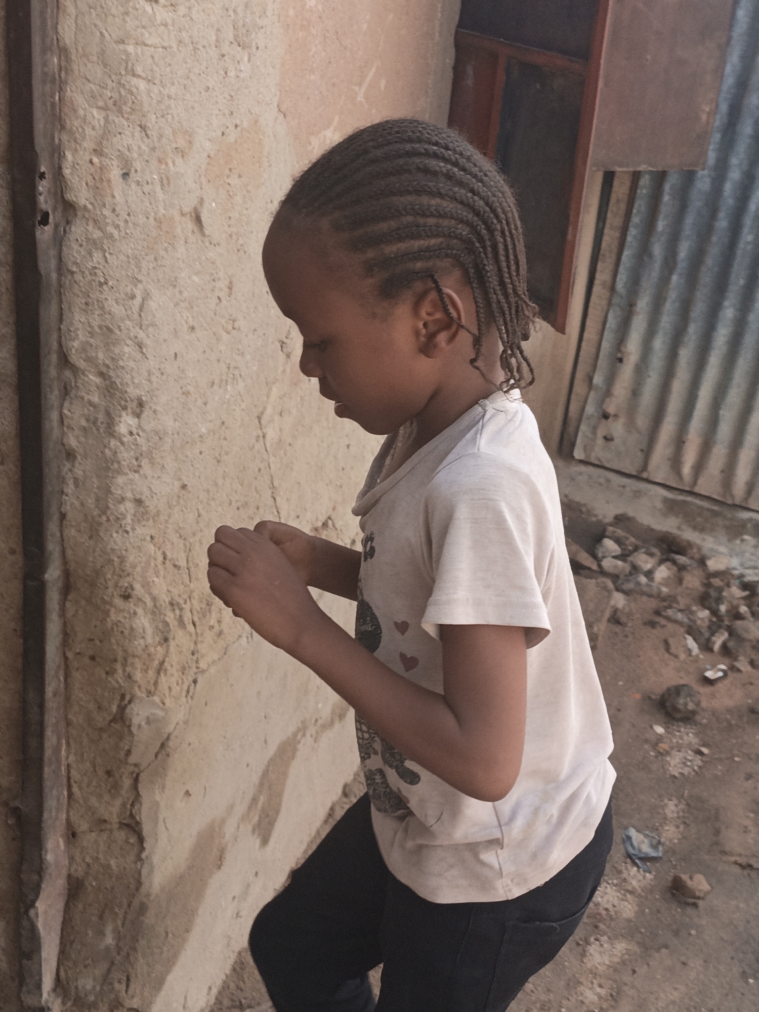The image shows a young girl in profile, looking down at her hands. She has braided hair and is wearing a light-colored t-shirt with a graphic design, and dark pants. She is standing outdoors next to a textured, light-colored wall. To her right, there is a corrugated metal structure and debris on the ground. The scene appears to be an outdoor urban environment.