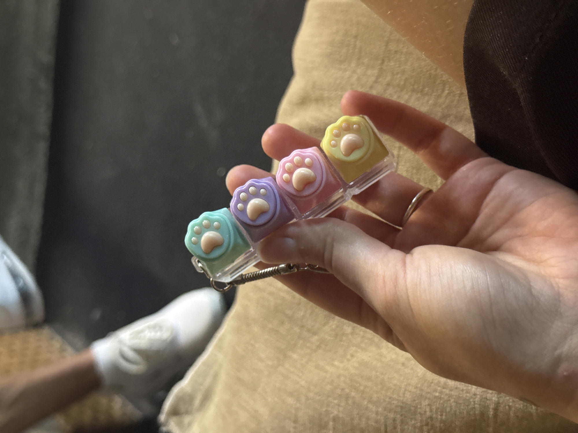 A hand is holding a small, colorful keychain with four paw-shaped charms. The charms are stacked vertically and are colored light blue, lavender, pink, and yellow, each with a lighter pink paw print. The keychain is attached to a silver spring. In the background, a person wearing white sneakers and dark pants is visible, with a textured, tan surface beneath. The setting appears to be indoors, possibly a living room or lounge area. There are no visible landmarks or contextual clues to suggest the location is Aksu, Turkey.