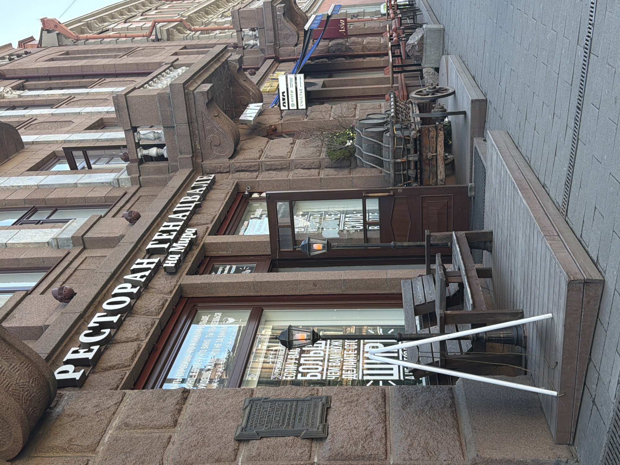 This media file depicts the street-level facade of a building, likely a commercial establishment, in a city. The scene captures the exterior of a restaurant during what appears to be daytime, possibly under an overcast sky, given the soft, diffused light and lack of strong shadows.
The main subject is a restaurant named "РЕСТОРАН ГЕНАЦВАЛИ на Мира" (Restaurant Genatsvali on Mira), displayed in prominent white lettering on the building's rough-hewn stone facade. The architecture features decorative elements such as stone cornices, ornate carvings, and several windows. The restaurant's exterior includes a wooden outdoor seating area with benches and tables, along with rustic decorative elements like old wooden barrels, a large wooden wheel, and antique-style iron lanterns. A small plant pot containing greenery is visible. Another sign, "Asia," is mounted above the restaurant's entrance area, suggesting another establishment or a diverse menu.
Notable details include various texts displayed on the windows and a metal plaque. One window features a poetic quote in Russian: "БЫТЬ СПРОСИШЬ, ЧТО Я ЛЮБЛЮ БОЛЬШЕ, ВИНО, Я ОТВЕЧУ ЧТО ВИНО, ТЫ ОБИДЕШЬСЯ УЙДЕШЬ, ЖАЛЬ, ЧТО НЕ ЗА ВИНОМ" (If you ask what I love more, wine, I will answer that wine, you will be offended, you will leave, it's a pity, not because of wine). Another section of text reads "ЕСЛИ У ВАС БОЛЬШЕ ЧЕМ ВАМ НУЖНО ДЕЛАЙТЕ СТО" (If you have more than you need, make a hundred). Smaller signs, "WAW," "КРУГИ" (Circles), and "СЧАСТЬЕ" (Happiness), are visible on an upper window. A historical-looking metal plaque on the wall states "ГРУЗИНСКАЯ ПОСОЛЬС" (Georgian Embassy), likely a playful reference to the restaurant's Georgian cuisine.
No people are visible in the image, conveying a quiet and static moment on the street. Based on the provided context, this scene is located in Krasnoyarsk, Russia.