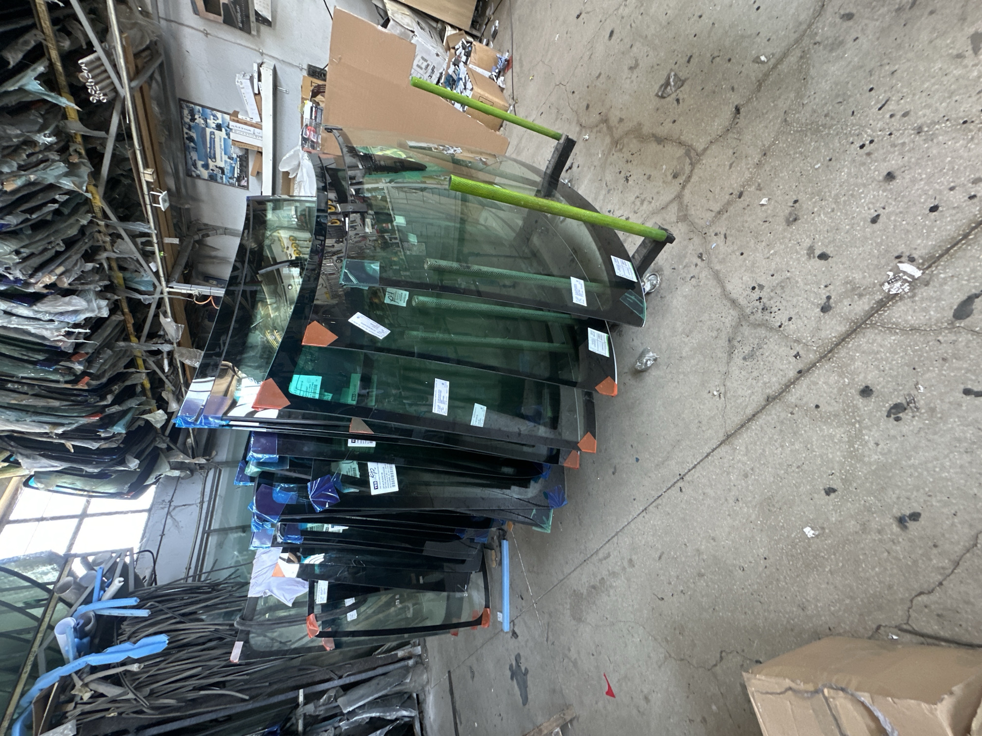 The image shows an indoor automotive salvage yard or glass shop. In the foreground, a row of windshields, presumably for cars, is arranged vertically, leaning against a rack. The windshields are various sizes and shapes, some with orange protective corner coverings. A metal rack with green cylindrical supports is holding some of the windshields. The floor is concrete, showing signs of wear and some scattered debris. In the background, more automotive parts, including what appear to be long rubber seals, are stored. Shelving units are visible further back, with boxes and other items stacked on them. A calendar is hanging on the wall, and a framed picture depicting cars is also present. The overall impression is of a storage or service area for automotive replacement parts.