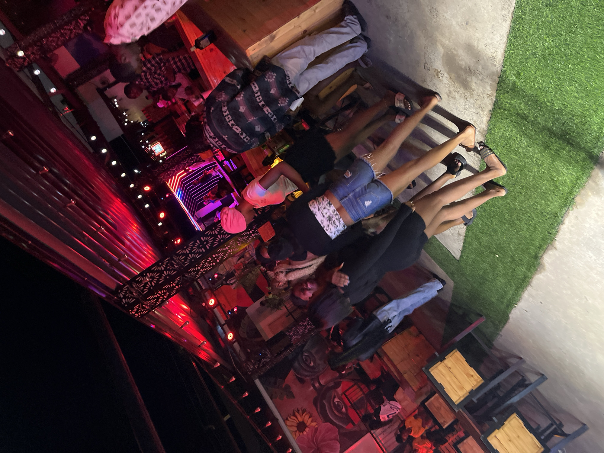 The image depicts a nighttime scene at an outdoor establishment in Howayi, Nigeria, likely a bar or lounge. Red and pink lighting illuminates the space, reflecting off the corrugated metal ceiling structure. Several individuals are present, most of whom are young adults.
In the foreground, a group of young women is standing together. One woman on the left wears a black top and jeans. Next to her, another woman has a white patterned top and denim shorts. To her right, a woman wearing a pink headscarf and a light-colored top stands with a black skirt. Another woman, with dark hair, is partially obscured behind them.
In the midground, a man in a patterned shirt and light-colored pants is visible. Further back, other patrons are seated at tables, and some are standing. The area features outdoor seating with wooden tables and chairs, and a patch of artificial grass. A mural adorns one of the walls, featuring stylized facial features and flowers. The overall atmosphere suggests a lively social gathering.