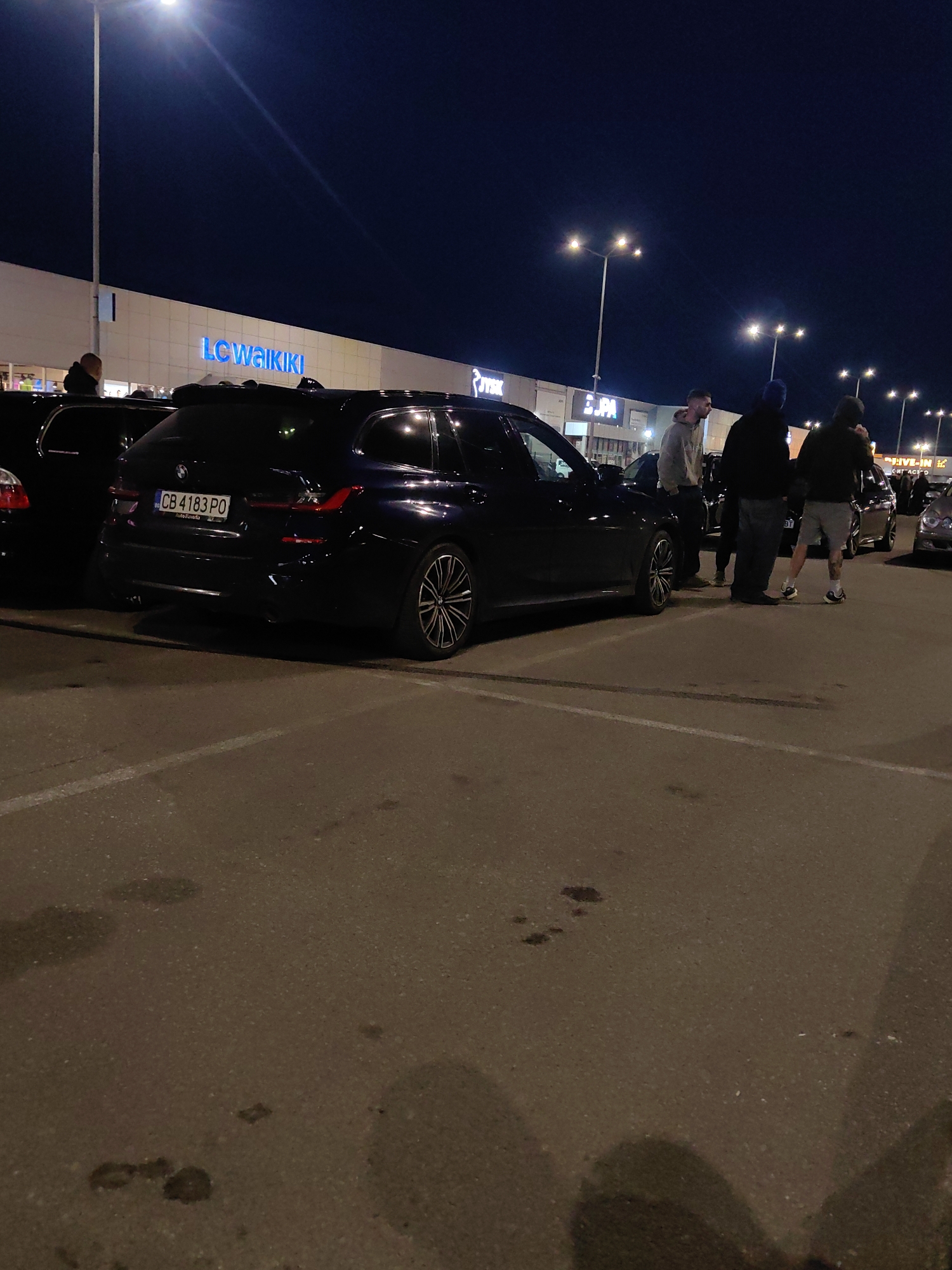 The image captures a night scene in what appears to be a parking lot, possibly near a commercial area. In the background, there is a large building with the 'LC WAIKIKI' signage visible. Several other signs, including 'JYSK' and 'JPA', are also seen on the building. A dark-colored BMW wagon is parked and the Bulgarian license plate 'CB 4183 PO' is visible on the car. Nearby, a small group of people are standing next to the vehicle and interacting. Streetlights provide illumination. The pavement shows some damp spots. The location appears to be in Bulgaria, based on the license plate style, and possibly in the city of Sofia if the ‘CB’ prefix is considered.