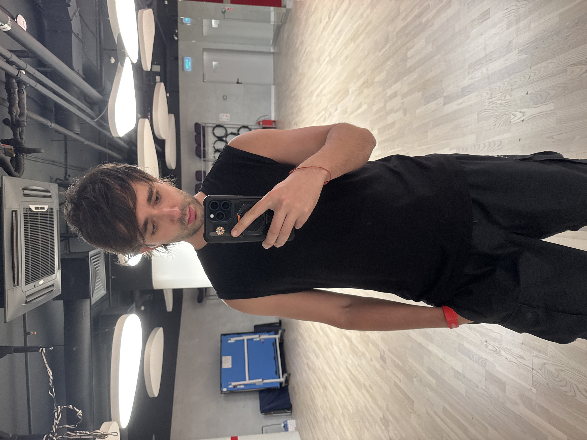 A young man is taking a mirror selfie in what appears to be a dance studio or gym. He is wearing a black sleeveless shirt and black cargo pants. He has dark hair and is looking down at his phone. He is wearing a red wristband on his left wrist and a thin red string bracelet on his right wrist. In the background, there is a blue ping pong table, some exercise equipment, and large oval-shaped lights on the ceiling. The floor is made of light-colored wood. The overall environment suggests a modern, well-lit indoor space. There is no indication of the location being Moscow, Russia.