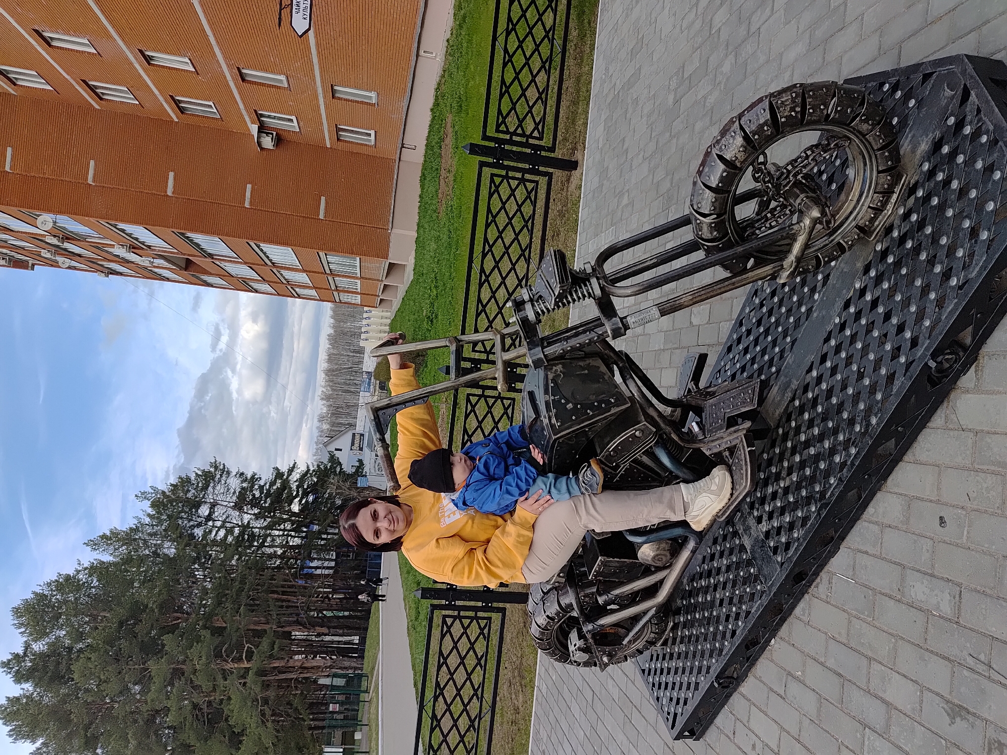 This outdoor photograph, taken in Chaikovsky, Russia, captures a woman and a child sitting on a large, dark metal motorcycle sculpture. The image is framed to show a multi-story brick building, a black metal fence, and a line of tall pine trees in the background under a partly cloudy blue sky, suggesting daytime.
The primary subjects are a woman, smiling brightly, wearing a yellow sweatshirt with "Be the" visible text, light-colored pants, and white sneakers. She is holding a young child, likely a toddler, dressed in a blue jacket and a black beanie, on her lap. The child appears content. They are both seated on an intricately designed, industrial-style motorcycle sculpture, crafted from dark metal, featuring a prominent, rugged front wheel with studded details and chains.
The sculpture rests on a black metal diamond-plate platform, which in turn sits on light gray brick pavers. Behind the fence, green grass is visible. In the distance, a blurry sign on a building reads "Запчасти" (Spare parts). On the closer brick building, a white sign with black text is partially visible, showing "НАЦИОНАЛЬНЫЕ КУЛЬТУРЫ" (National Cultures). The overall scene depicts a pleasant, casual moment of enjoyment in what appears to be a public area or park.
