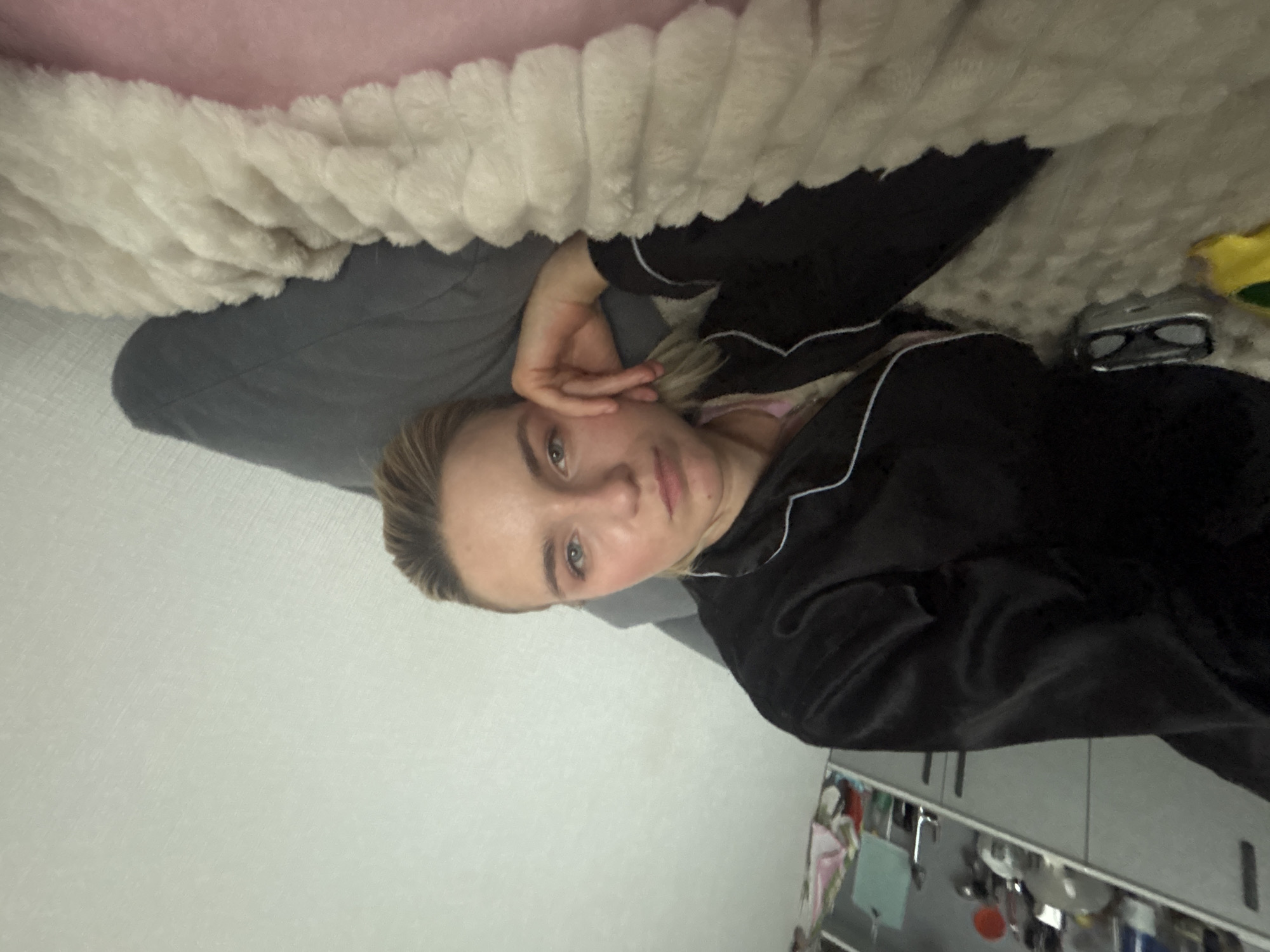 A woman lies on a bed under a blanket. She is wearing black pajamas with white piping. The bed has a pink headboard, a gray sheet, and a white furry blanket. Glasses are on the bed to her right. A shelf or cabinet is visible in the background with various objects on it, suggesting a domestic interior. The location is Unknown, Russia, based on the prompt.