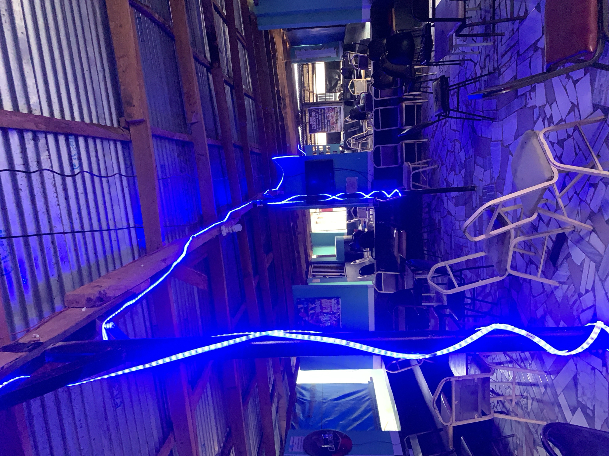The image depicts an indoor space in the New Stadium area of Nigeria, characterized by corrugated metal walls supported by wooden beams, illuminated by blue LED strip lights. The ceiling is low. The floor has a mosaic pattern. The room contains tables and chairs, some of which are stacked. There are posters or signs visible on the wall. The overall lighting is low and creates a blue cast. There are no people visible in the image.