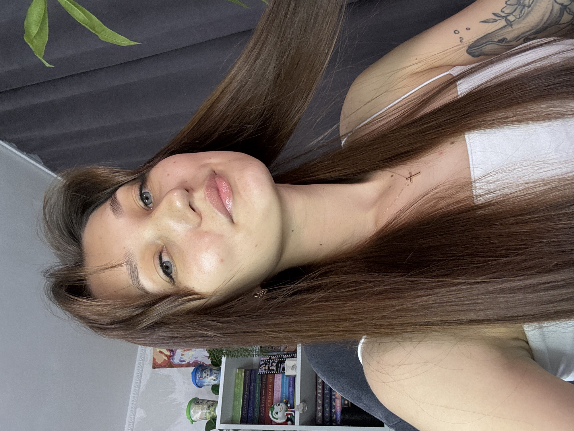 A young woman with long, straight brown hair is positioned in a close-up shot, looking towards the viewer. Her skin has a light complexion with a scattering of freckles across her nose and cheeks. She is wearing a white tank top with thin straps, and a delicate gold cross necklace is visible. Her hair is falling across her shoulders and partially obscuring her left side. In the background to her left, a white bookshelf is filled with books and small decorative items. To her right, a dark grey curtain hangs, and a portion of a green plant is visible. The lighting suggests an indoor setting, possibly a home, and there are no indications of specific locations within Novokuznetsk, Russia.
