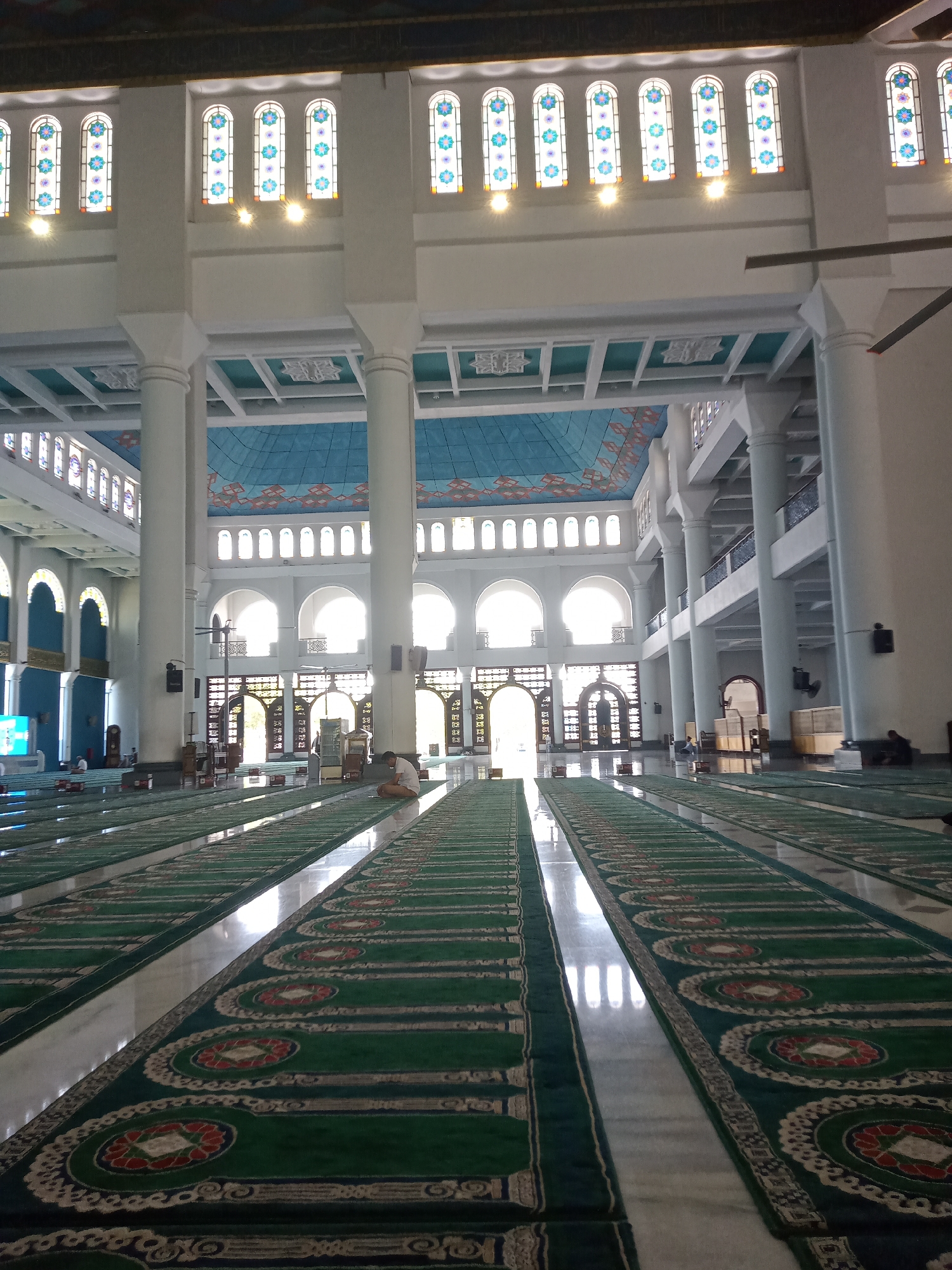 The image depicts the interior of the Al-Akbar National Mosque in Surabaya, Indonesia. The perspective is from the ground level, looking down the length of the prayer hall. Rows of green prayer mats with intricate white and red floral patterns are laid out on a polished marble floor, leading towards large arched doorways at the far end. Sunlight streams in from these doorways, creating strong reflections on the floor and illuminating the space.
Large, white cylindrical columns support the high, ornate ceiling, which features blue panels with geometric patterns and decorative elements. Arched windows with stained glass depicting floral motifs are visible along the upper walls, letting in diffused light. In the middle distance, a lone individual is seated on a prayer mat, facing away from the camera. Several other figures are sparsely scattered throughout the vast hall, some seated or standing. The overall impression is one of grandeur and tranquility.