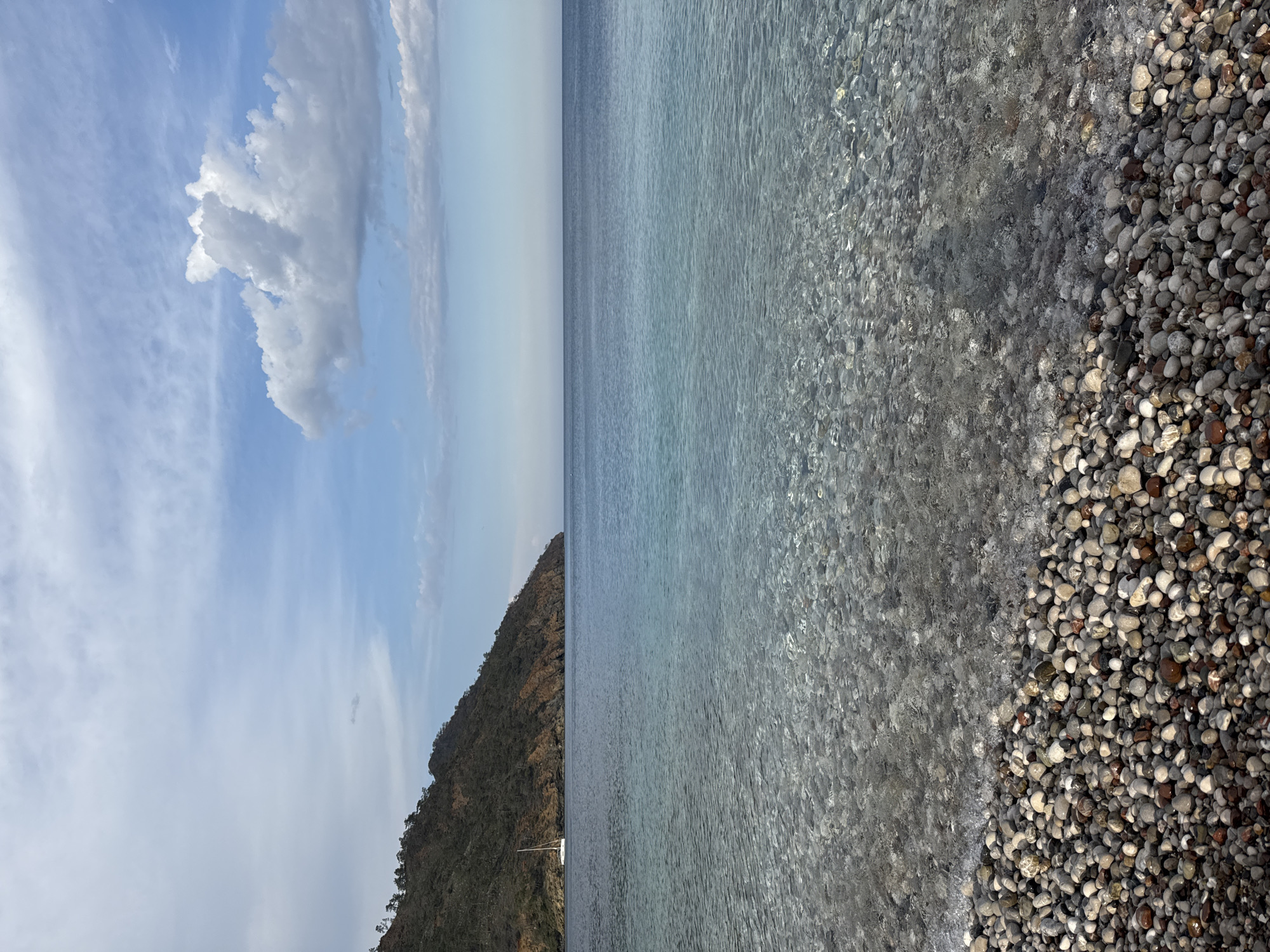 This serene landscape photograph captures a tranquil coastal scene in Kumluca, Turkey. The image, presented in a horizontal orientation, is divided horizontally into distinct sections. The top portion features a clear, bright blue sky adorned with scattered white, fluffy clouds, indicating fair weather.
The horizon line bisects the frame, separating the sky from the expansive, calm sea below. On the left side of the image, a dark, densely forested mountain or hill slopes down, meeting the clear waters of the sea. A small, white sailboat is visible, anchored peacefully near the base of this mountainous coastline, its mast slender against the dark land.
The right half of the image showcases the crystalline water, which is so clear that the diverse array of pebbles on the seafloor is distinctly visible. These smooth, rounded stones, in varying shades of grey, brown, and white, transition from submerged to dry, forming a natural pebble beach along the extreme right edge of the frame. The gentle ripples on the water's surface reflect the bright daylight.
The scene conveys a sense of peaceful natural beauty, highlighting the clear waters, the rugged coastline, and the serene atmosphere of a sunny day by the sea. No people are visible, and there is no discernible text in the photograph. The time of day appears to be daytime, possibly late morning or early afternoon, given the bright illumination.