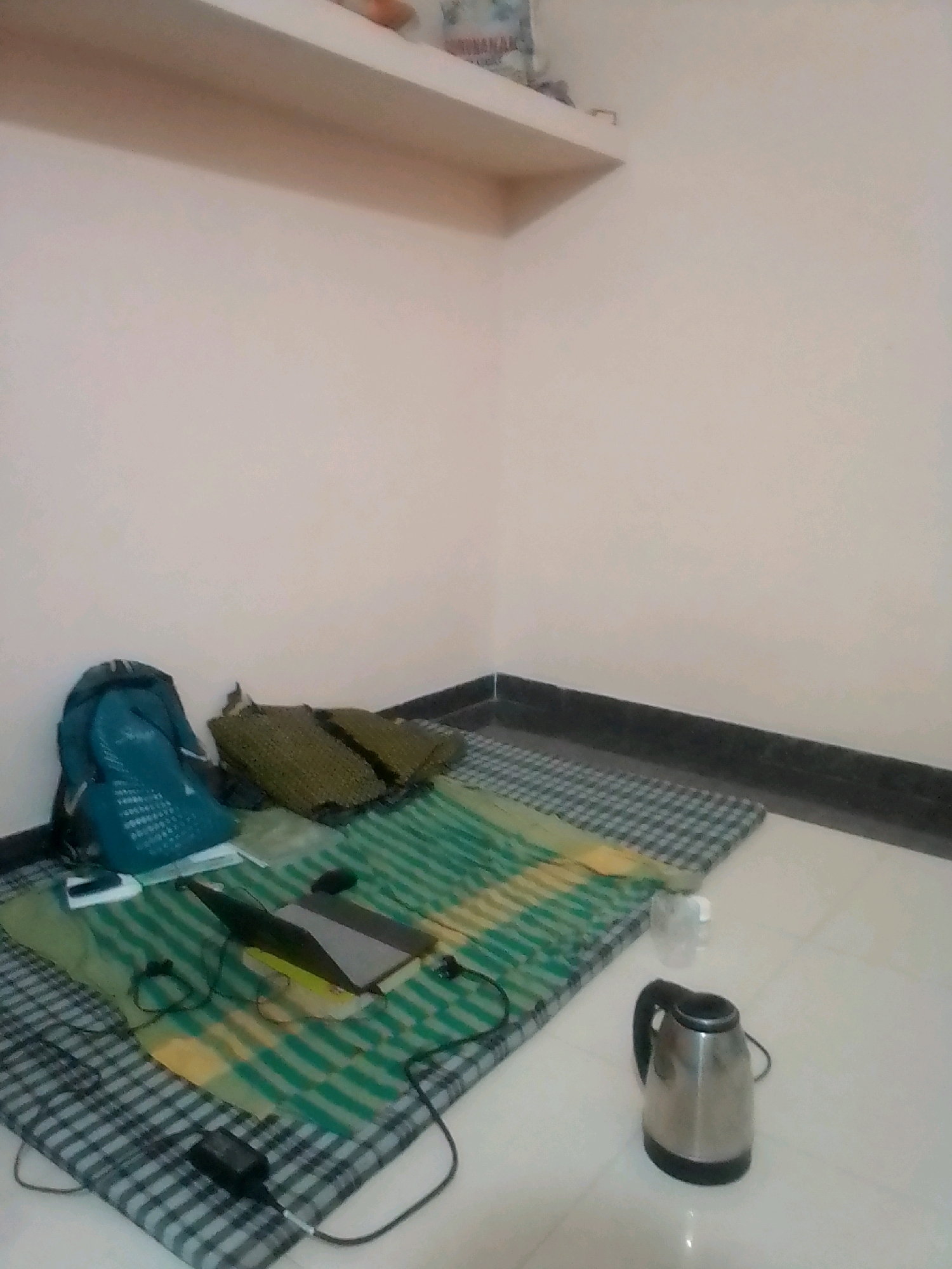 The image depicts a simple, minimalist living space, likely a bedroom or a study area, within a residence in Anjaniya, India. The floor is covered with a checkered mattress, upon which a green and yellow striped sheet is laid out. To the left, a teal backpack rests against the wall, next to two woven mats. In the center of the laid-out bedding, a laptop is open, with wires and a power adapter visible, suggesting a workspace. A small white object, possibly a mobile phone or a portable charger, is also present. A glass of water stands to the right of the laptop, and a stainless steel electric kettle is placed on the floor further to the right. The walls are painted a light, neutral color, and a thin shelf is mounted high on the wall. The overall impression is one of a functional and uncluttered personal space.