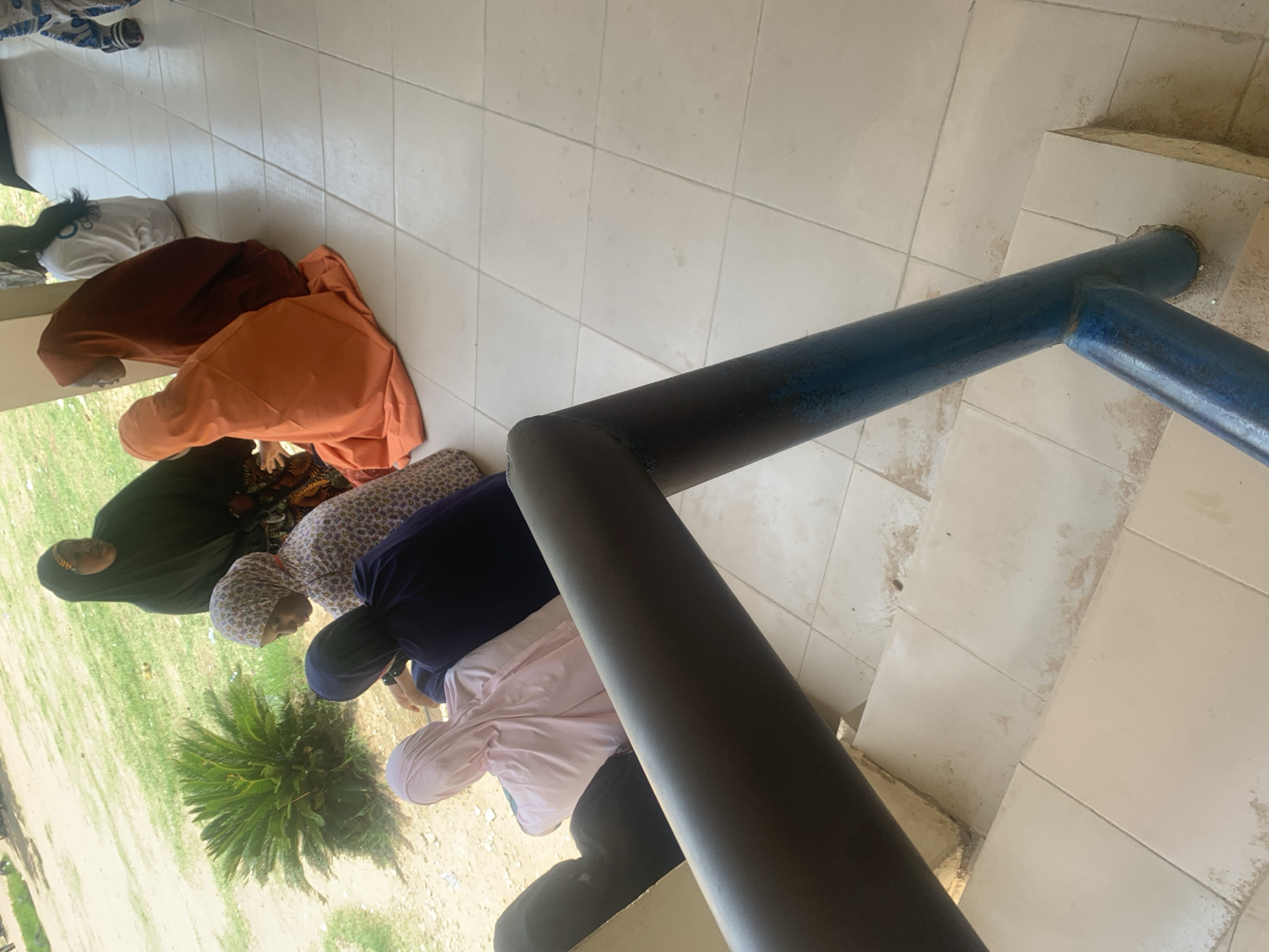 The image shows a group of women, dressed in hijabs and modest attire, kneeling on a tiled floor. They appear to be engaged in prayer, with their heads bowed. The setting seems to be outdoors, with a glimpse of green grass and dirt visible to the left of the frame. A dark blue metal railing runs diagonally across the image, partially obscuring the women. The tiles on the floor are light-colored and appear somewhat worn or stained. The overall lighting suggests it could be daytime, possibly morning or afternoon, with no clear indication of weather conditions. The orientation of the image is landscape, and it has been rotated, so the people appear to be on an incline. There is no visible text in the image. The context provided places the scene in Jalingo, Nigeria.
