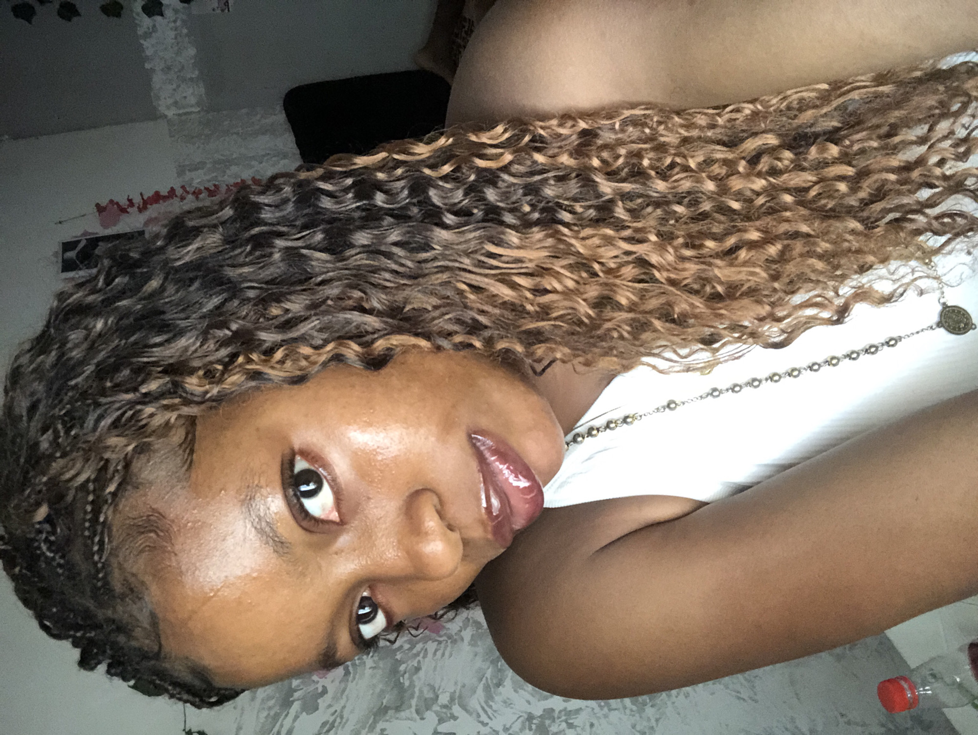 A close-up, portrait-style photograph depicts a young, dark-skinned woman with partially braided and wavy hair. Her hair transitions from dark at the roots to light brown and blonde at the ends. She is wearing a white top and a silver necklace. Her lips appear glossy.
The setting is indoors, with a patterned wall visible in the background. A black object, possibly a piece of furniture, is also partially visible in the background. A water bottle is in the bottom right corner of the image. The location is Unknown, Nigeria.