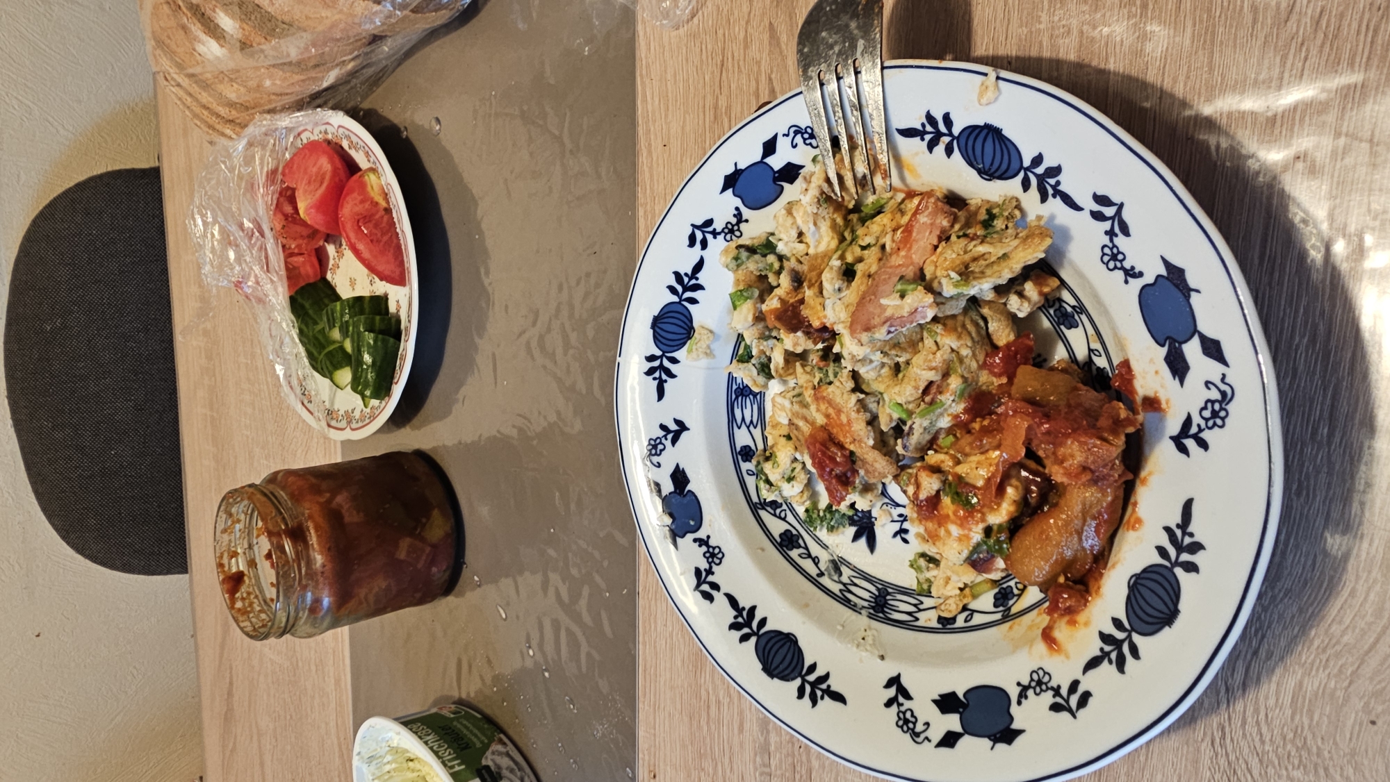 The image shows a top-down view of a meal laid out on a wooden table. The primary subject is a plate of scrambled eggs, mixed with what appears to be pieces of sausage or ham, and garnished with chopped green onions. The eggs are served with a portion of what looks like a tomato-based sauce or relish. A fork is placed on the plate, indicating the food is ready to be eaten.
To the left of the plate, there is a small white dish containing sliced tomatoes and cucumber. Adjacent to this dish is a glass jar, partially filled with a reddish, chunky substance, likely a condiment or spread. Further left, a dark, textured surface, possibly a chair or cushion, is visible. In the bottom right corner, part of a white container with visible text is peeking into the frame. The text appears to say "Frischkäse," which translates to "cream cheese" in German.
The overall setting suggests a casual meal, possibly breakfast or lunch, in a domestic environment. The lighting suggests it could be daytime, with shadows cast on the table. There are no people visible in the image. The scene is set in Oelsberg, Germany.