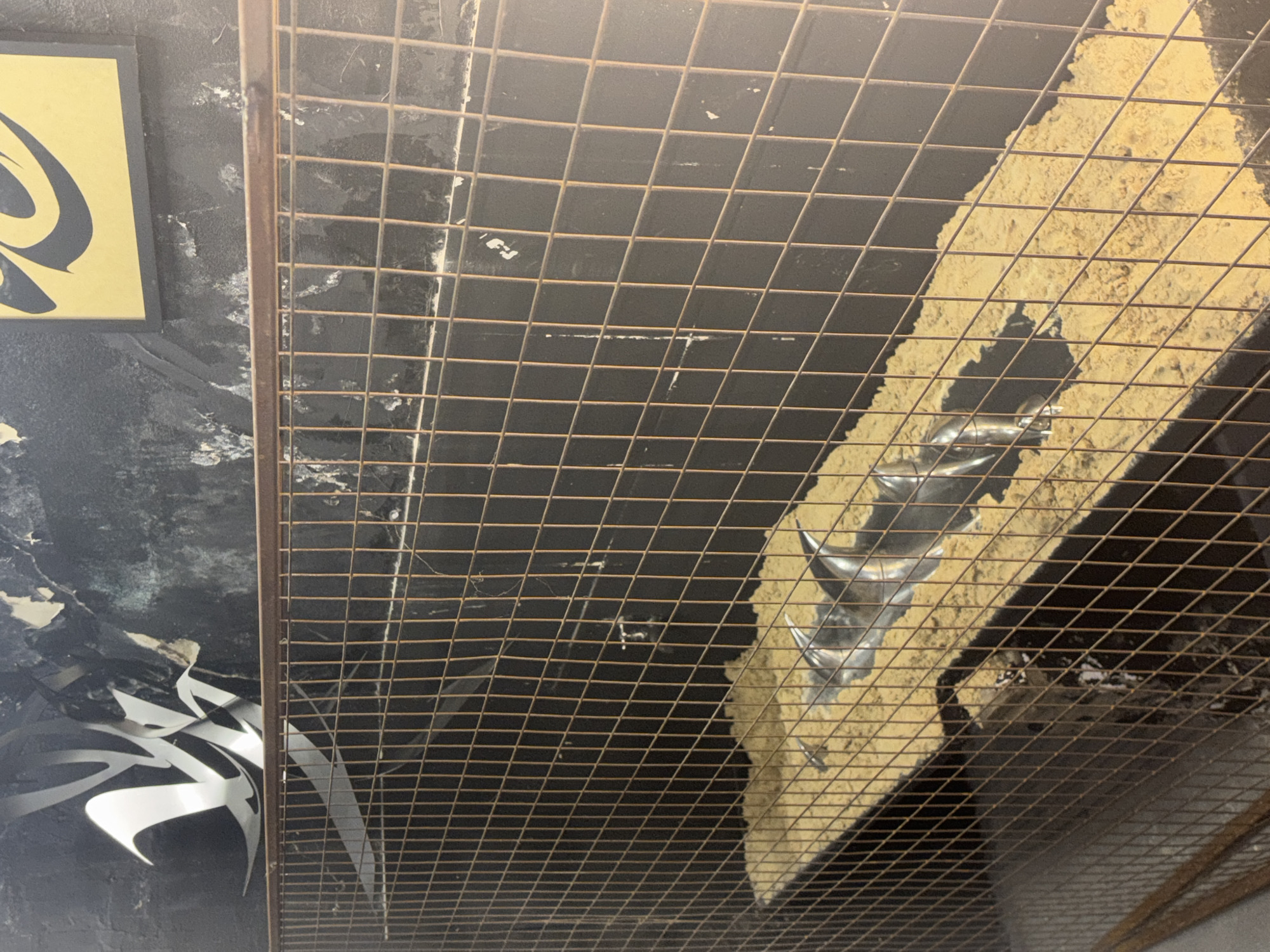 The image shows a metal grid structure obscuring a view of an interior space. On the left, a dark wall with black and white graffiti-like designs is visible, along with a yellow and black rectangular sign. Through the grid, a diagonal section with a tan, possibly textured surface is seen, featuring a metallic, corkscrew-shaped object partially embedded within it. The lighting is uneven, with shadows cast by the grid. The location in the city of Buenos Aires, Argentina, is unknown based solely on the visual information provided.