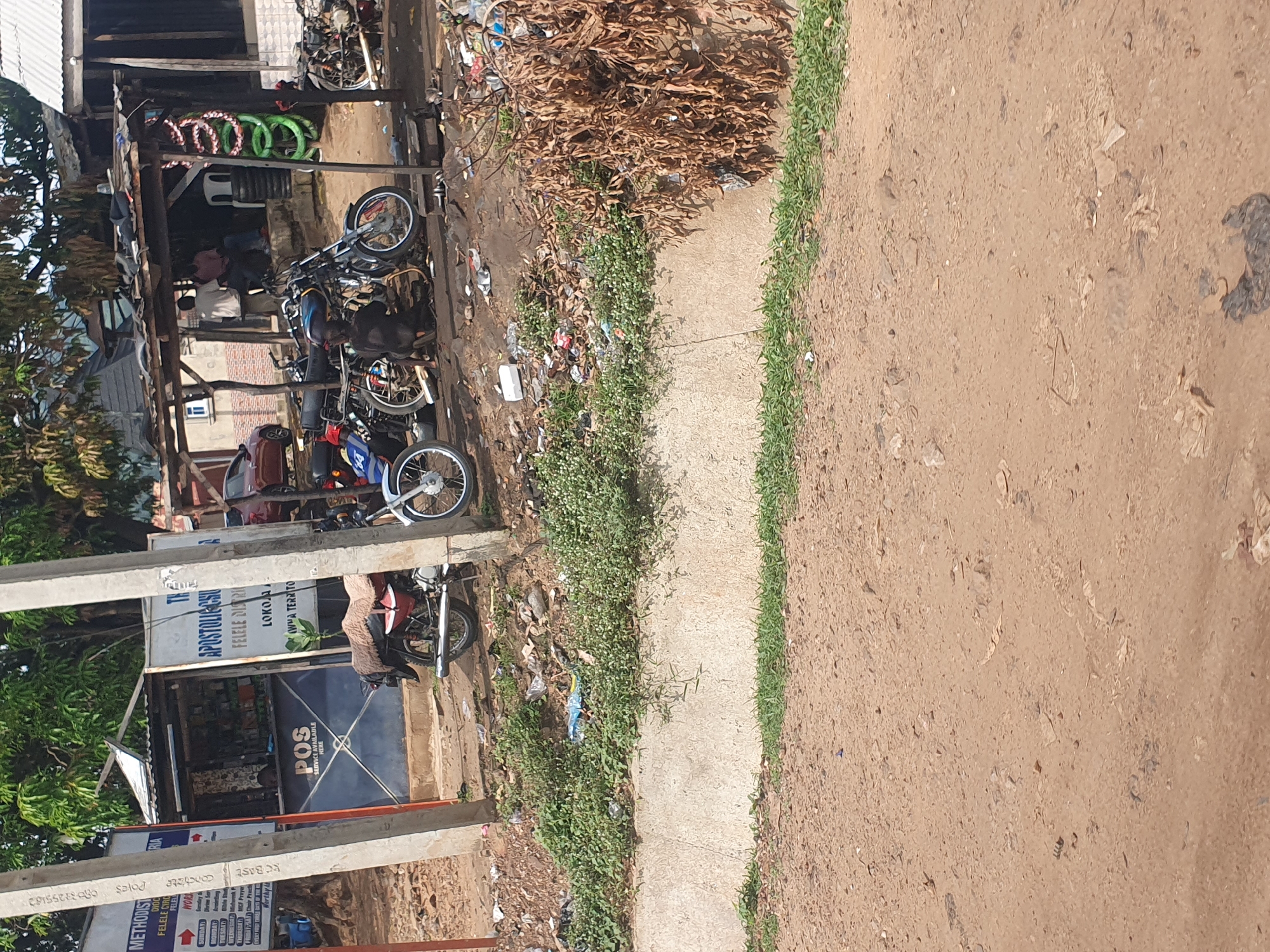 The image depicts a street scene in Lokoja, Nigeria, near a roadside establishment. In the foreground, a dirt road is visible, with a concrete drainage channel running horizontally across the middle ground. Beyond the drainage, there is a collection of motorcycles parked and undergoing maintenance. One individual, appearing to be a mechanic, is crouched down working on a motorcycle. A few other people are seated under a makeshift shelter, observing. Various items, including tires, spare parts, and debris, are scattered around the work area. A signpost for "APOSTOLIC CHURCH FELELE DISTRICT" and "POS SERVICE AVAILABLE HERE" is prominently displayed on a building. The overall environment appears to be a bustling urban area, typical of roadside services and commerce in Nigeria.