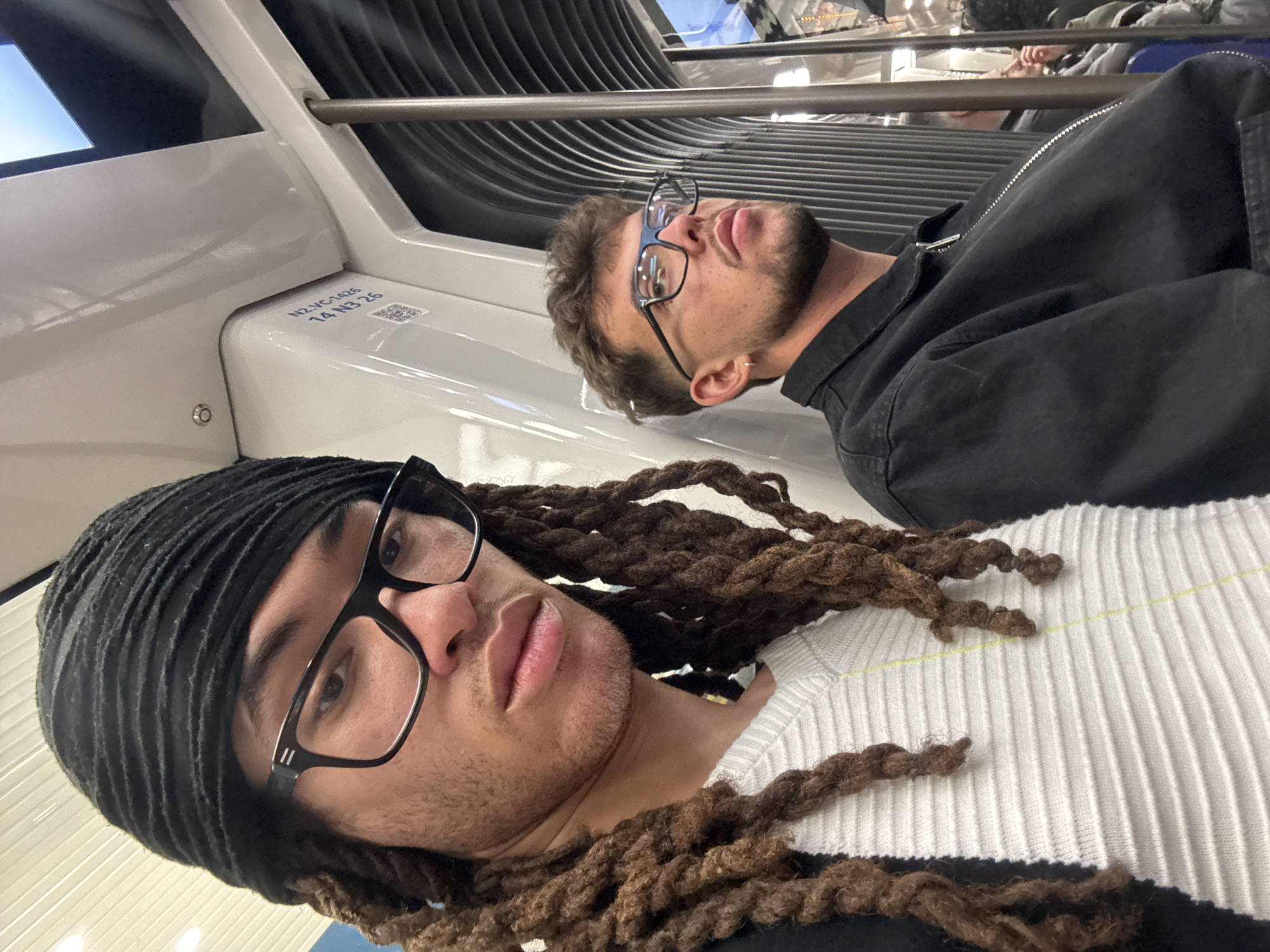 This media file captures two individuals inside what appears to be a modern public transport vehicle, likely a metro or train in Paris, France. The image is rotated 90 degrees clockwise, presenting the subjects horizontally.
In the foreground, closer to the camera, is a person with dreadlocks, wearing a black head wrap and black-framed glasses. They have light facial hair and are wearing a white ribbed sweater. Their gaze is directed towards the camera in a selfie-style shot, with a neutral expression.
Behind and to the right of the foreground subject, the second individual is visible. They have short brown hair, a beard, and are also wearing black-framed glasses. This person is dressed in a black jacket and is looking upwards or away from the camera, appearing contemplative.
The setting is an interior of a vehicle, characterized by white, curved panels that form seating or part of the carriage structure. A silver metal handrail runs along the top section. The background features ribbed, dark-colored panels, possibly part of the ceiling or a partition, and reflections from windows or screens are visible.
A notable detail is a label on a white panel to the left of the background subject. The text "N2-VG-1425" and "14 N3 26" is visible, along with a small QR code. The clean, modern design of the interior suggests a contemporary public transit system, consistent with parts of the Parisian metro or RER. The individuals appear to be commuters or passengers, with no specific activity other than being present in the vehicle. The lighting suggests daytime.