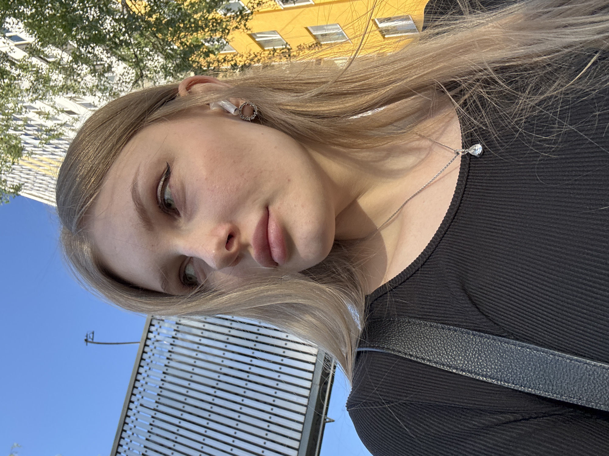 A young woman with long blonde hair, wearing a black top and a silver necklace, is captured in a selfie. She has fair skin, full lips, and wears subtle eye makeup. An earbud is visible in her left ear. The background reveals an urban environment with a blue sky, a modern architectural structure, and glimpses of a yellow building and green trees. The location is in Perm, Russia.