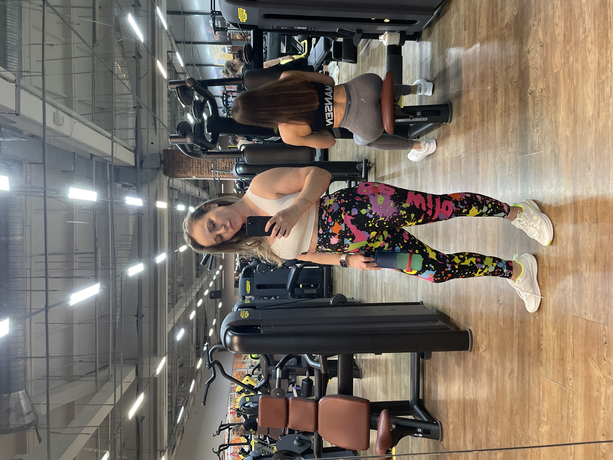 **Scene:** Gym interior, Chaikovsky, Russia.
**People:** Two women are present. One woman is holding a smartphone, taking a selfie, and wearing multicolored leggings and a white top. The other woman has brown hair, wears a black sports bra with white text, and grey leggings, and is using a seated leg curl machine.
**Objects:** Gym equipment including leg curl machines, weight machines, dumbbells, a smartphone, and the women's workout attire.
**Actions:** One woman is taking a selfie. The other woman is using a leg curl machine.
**Environment:** The gym has a wooden floor, a high ceiling with exposed metal beams and lighting fixtures, and rows of exercise machines.
