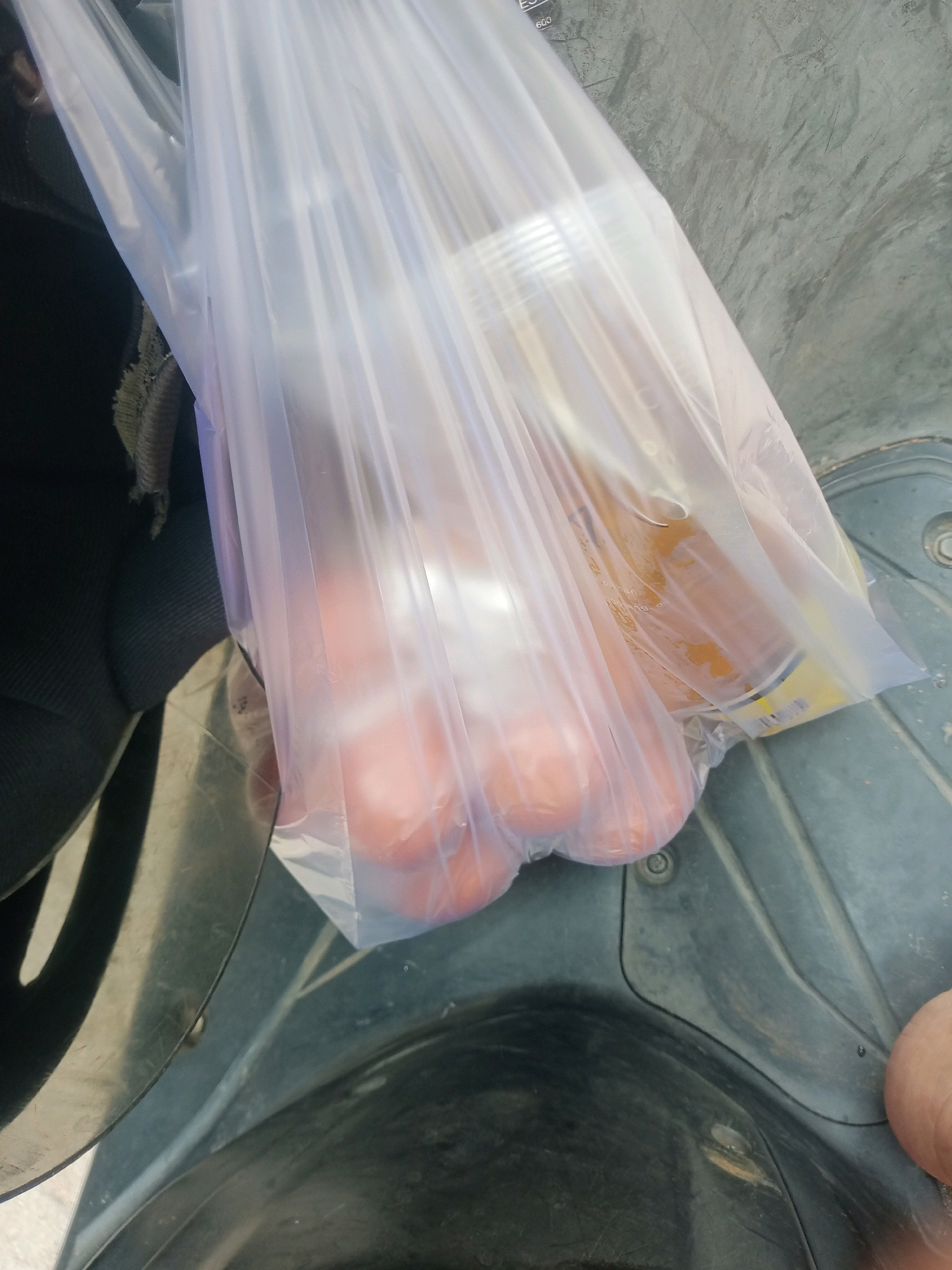 A plastic bag containing several round, red objects and a yellow can is positioned on a dark grey, textured surface, possibly the floorboard of a vehicle. The objects inside the bag appear to be tomatoes. The environment suggests an outdoor setting, with a glimpse of what could be a scooter's interior or a similar vehicle. The overall lighting is bright, casting shadows and highlighting the translucent plastic bag. The surface displays some wear and tear, with faint scratches and dirt visible. The scene is captured from a high-angle perspective. No specific location in Bandar Lampung, Indonesia, can be identified from the image.