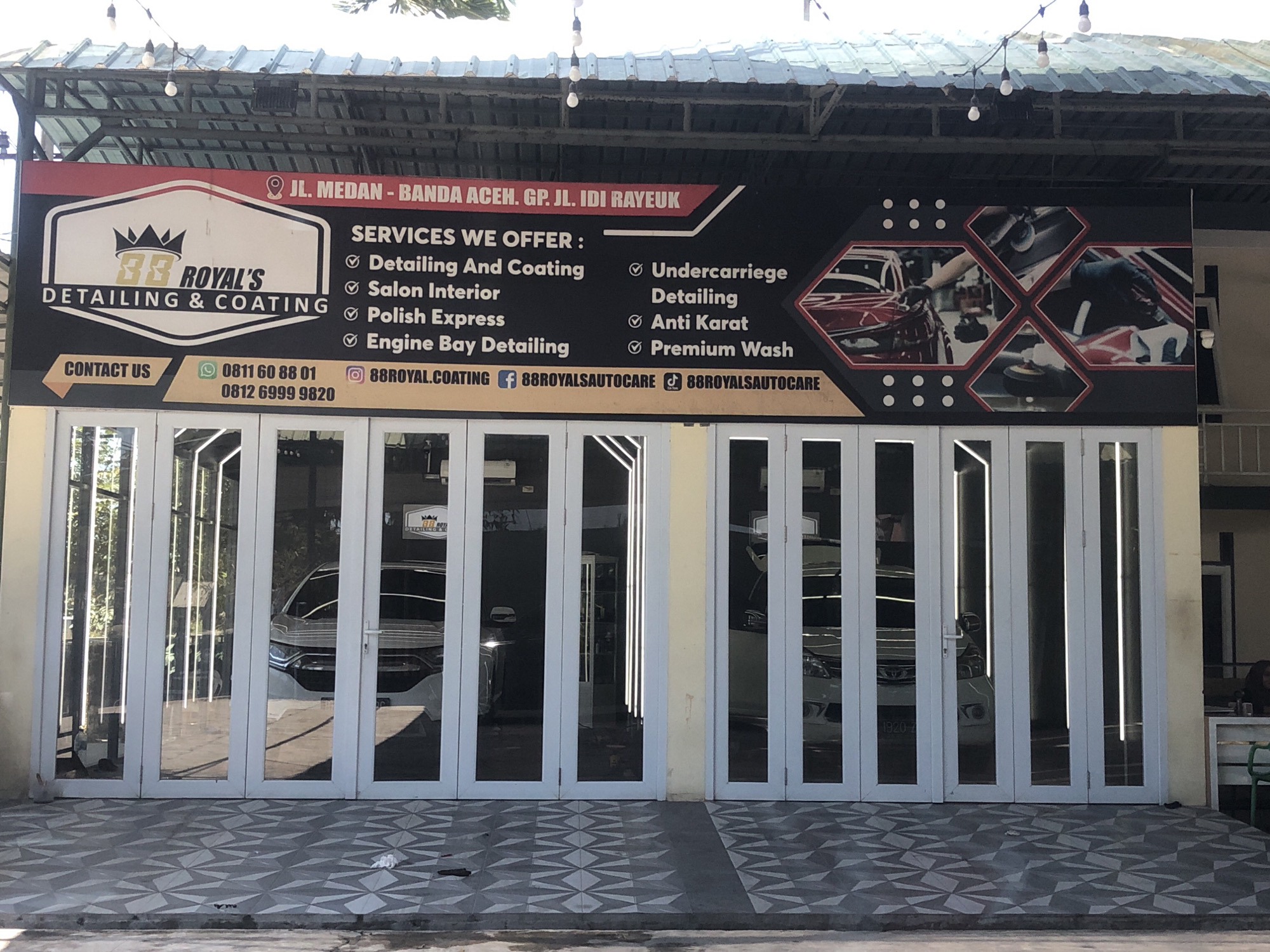 The image shows the exterior of a car detailing and coating business called "RR Royal's Detailing & Coating". The business is located on JL. Medan - Banda Aceh, GP. JL. Idi Rayeuk in Teupinbatee, Indonesia. The facade features a large banner with the business name, services offered, and contact information, including phone numbers and social media handles. Behind the banner are large, white bi-fold doors that open into the business, revealing several cars inside. The area in front of the business has a tiled floor and is a bright, sunny day, with light reflecting off the cars and the building.