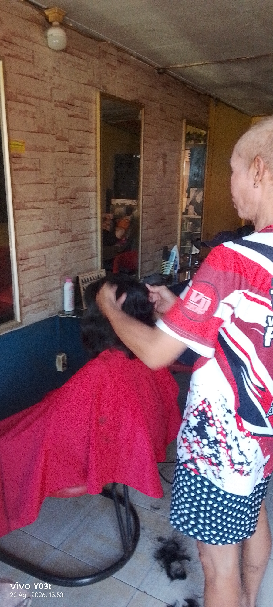 The image depicts a haircutting scene taking place outdoors in Dawidawi, Indonesia. A person with short, light-colored hair is cutting the dark hair of another person who is wearing a bright red cape. The backdrop includes a wall with a brick-like pattern and mirrors reflecting the surroundings. Scattered on the tiled floor are several small piles of cut hair. The overall impression is of a casual, informal haircutting service being offered.