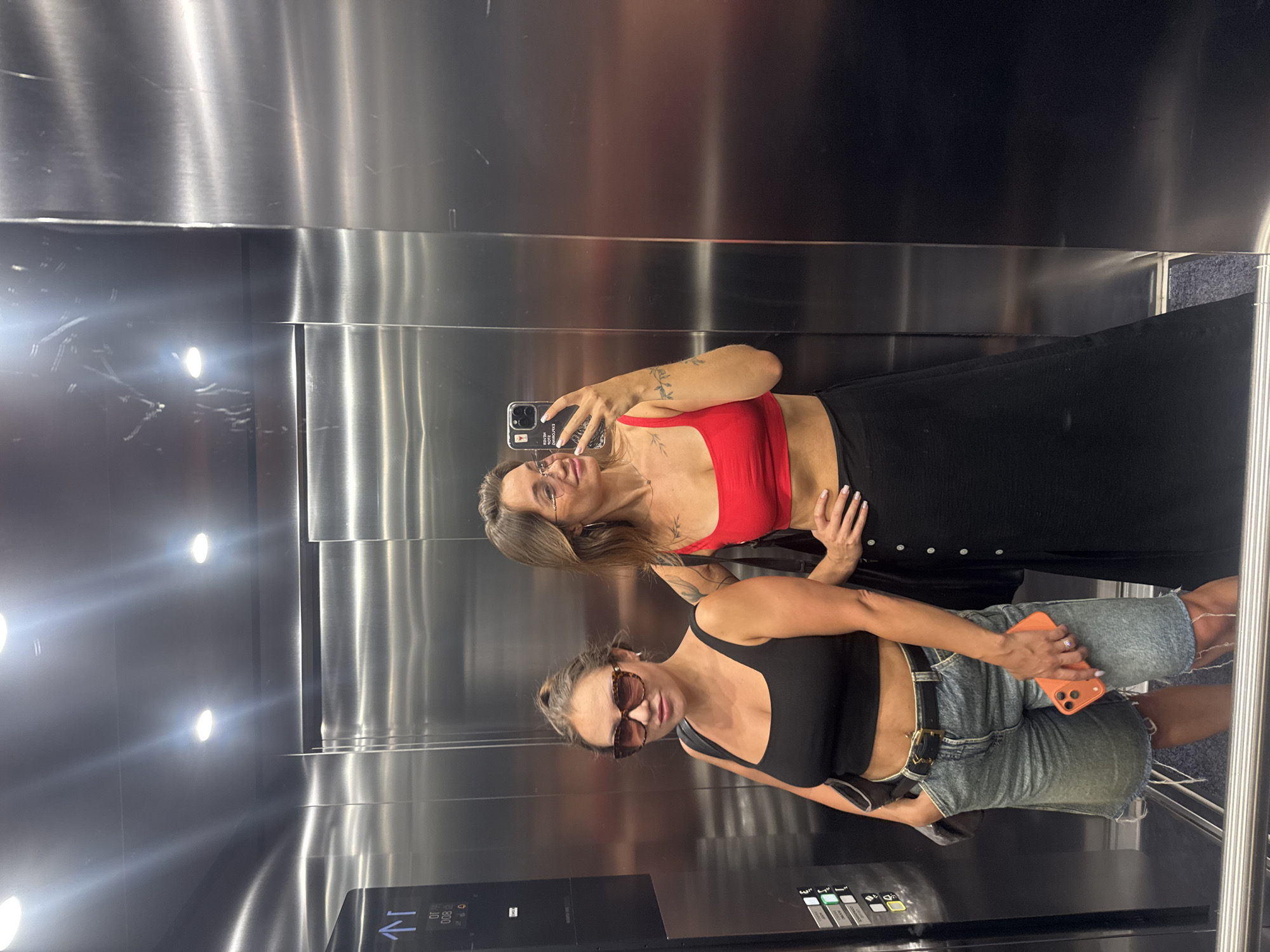 Two women are standing in an elevator with brushed metal walls. The woman on the left is wearing a black crop top, denim shorts, sunglasses, and is holding an orange iPhone. The woman on the right is wearing a red crop top, a long black skirt, and glasses. She is taking a selfie in the elevator mirror with her phone. The elevator appears to be in a building in Budva, Montenegro.