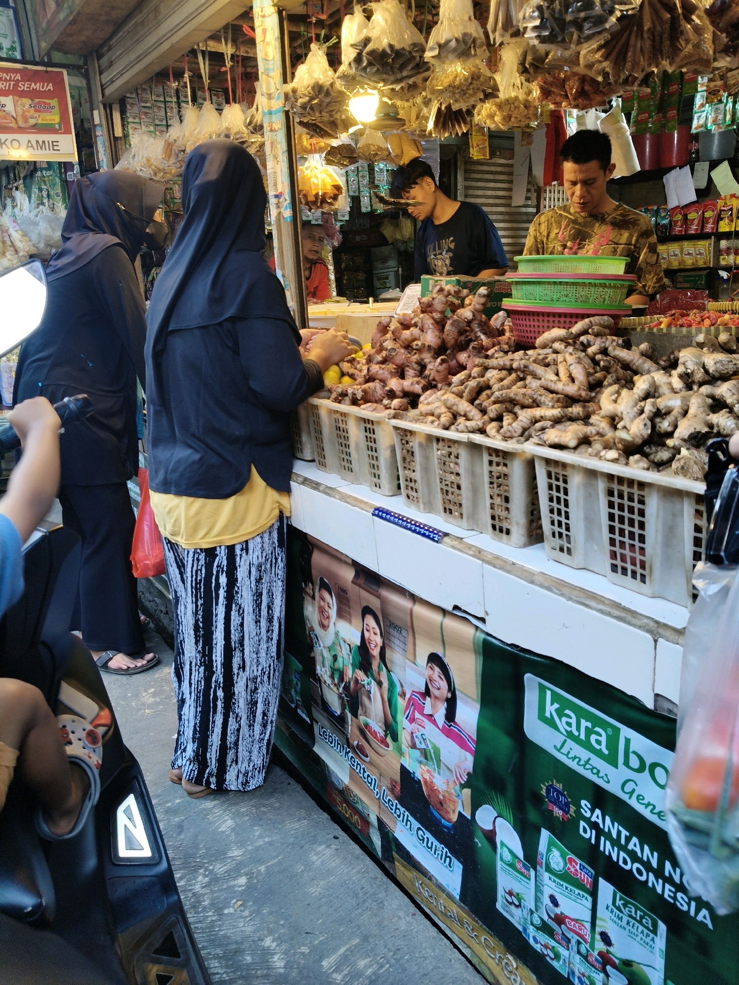 This is a medium shot, eye-level photograph taken outdoors, likely on a street market in Setiamekar, Indonesia. The scene depicts a bustling market stall filled with various produce and packaged goods.
In the foreground, on the left, a person's arm and leg are visible, with the leg wearing a colorful sandal. A portion of a dark-colored vehicle, possibly a scooter, is also present.
The central part of the image is dominated by a market stall. Three individuals are present: two women wearing hijabs and two men. One woman, wearing a black hijab and a black jacket over a yellow top and a black and white striped skirt, is seen from the back. She appears to be interacting with the stall owner. The second woman, also in a black hijab, is partially visible on the far left, facing away from the camera. One of the men, wearing a camouflage-patterned shirt, is standing behind the stall, looking down at the produce. The second man, wearing a dark blue t-shirt with white lettering, is also behind the stall, facing away from the camera and looking down.
The stall itself is laden with goods. Large quantities of ginger root are piled high in white plastic crates. Behind the ginger, there are stacks of green and pink plastic baskets. Further back, various packaged goods are visible on shelves, including small red bottles and other colorful items. Above the stall, an array of products in plastic bags are hanging from the ceiling. A large banner with Indonesian text and images of women and product packaging is displayed on the front of the stall.
The overall environment suggests an outdoor market setting with concrete flooring and a covered stall area. The lighting indicates it is daytime.