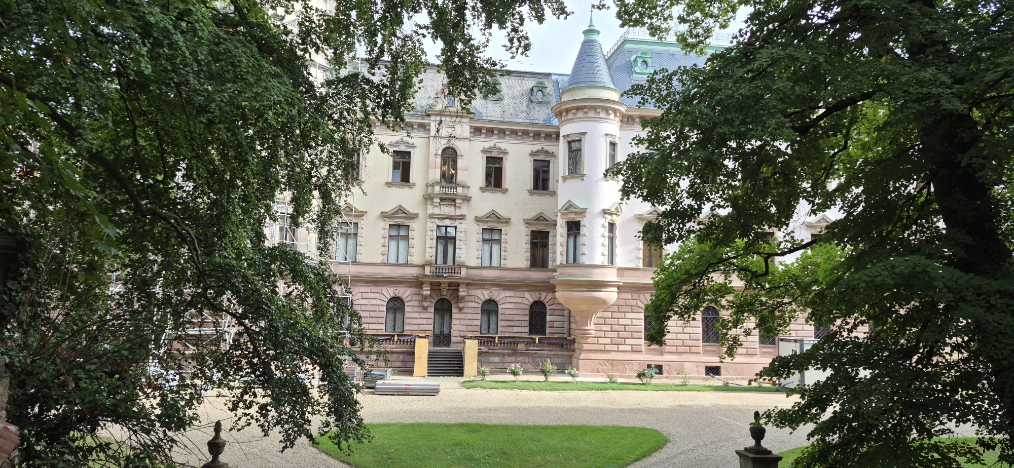 This image depicts the exterior of a historic building, likely a castle or manor, in Regensburg, Germany. The architecture is a blend of styles, featuring a light-colored facade with ornate detailing, arched windows, and a prominent conical tower with a grey slate roof. The building is partially obscured by lush green foliage from large trees in the foreground, suggesting a park-like setting. In front of the building, there is a gravel pathway leading to a central grassy area. Construction materials, including scaffolding and stacked planks, are visible near the entrance, indicating ongoing renovation or maintenance work. The sky is overcast, casting a soft light on the scene. There are no people visible in the image.