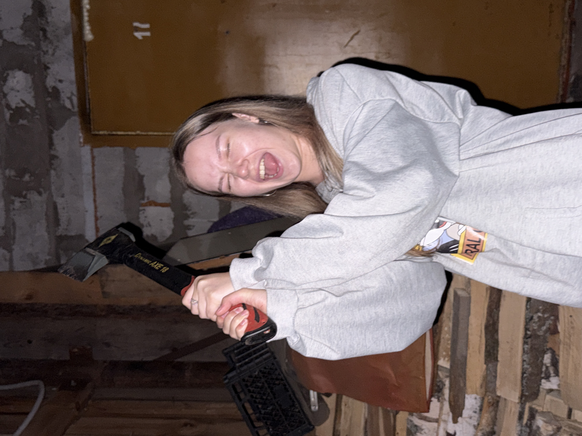 A young woman with blond hair is holding a "Dynamic AXE 18" axe in both hands. She is making a screaming face, with her eyes closed and mouth open. She is wearing a light grey hooded sweatshirt with a cartoon graphic on the sleeve and a ring on her left ring finger. The background features a brown wooden door with the number "17" on it, exposed brick, and stacks of firewood. A black plastic crate is also visible. The location is potentially Zhigalki, Russia.