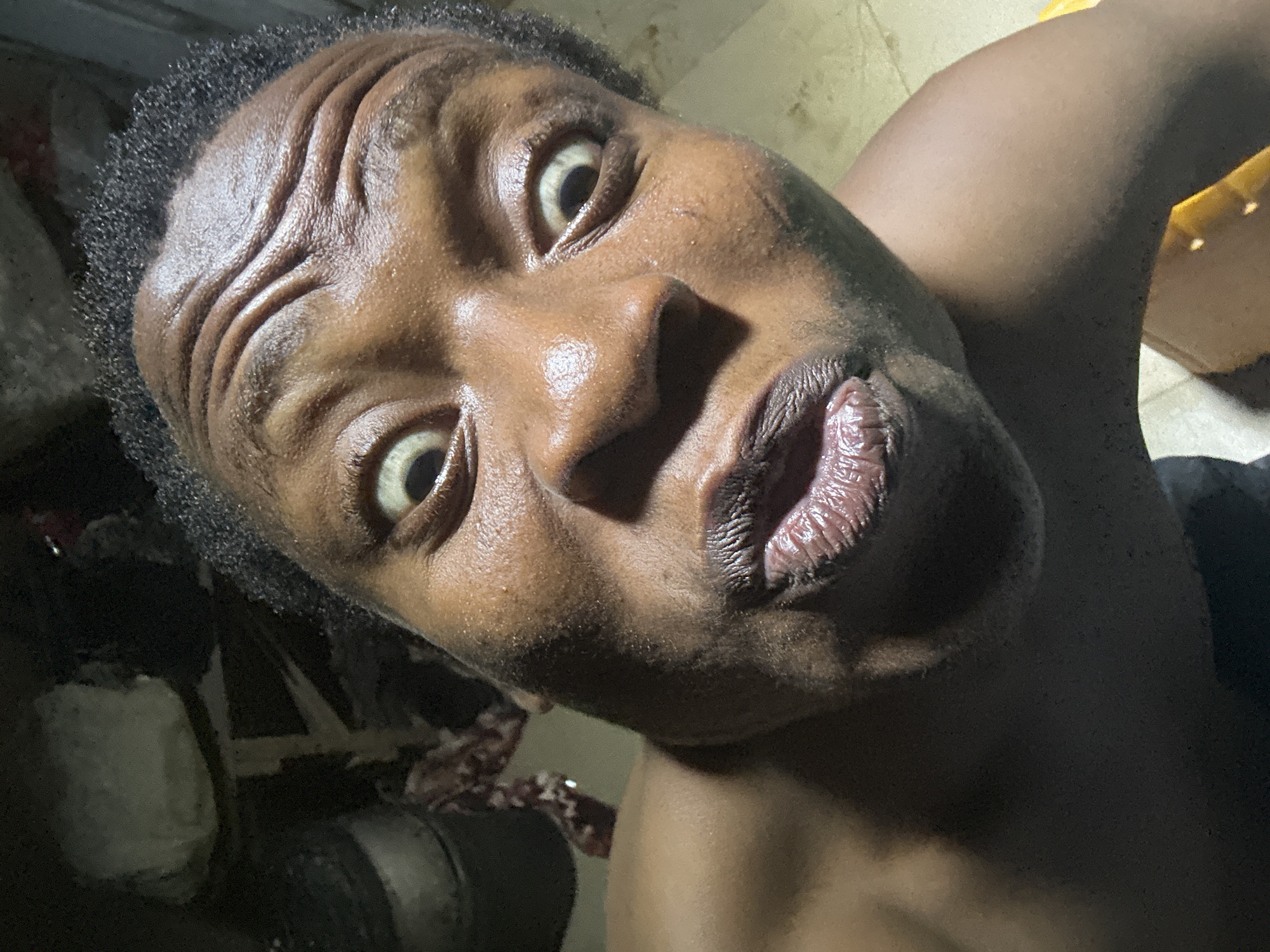 A close-up, high-angle shot of a Black man's face. He has dark, curly hair and wide, expressive eyes. His forehead is furrowed with wrinkles. The man's lips are pursed, giving a pouting expression. The background is dark and indistinct, with a few objects barely visible. The lighting is uneven, creating shadows on the man's face. No city information is provided.