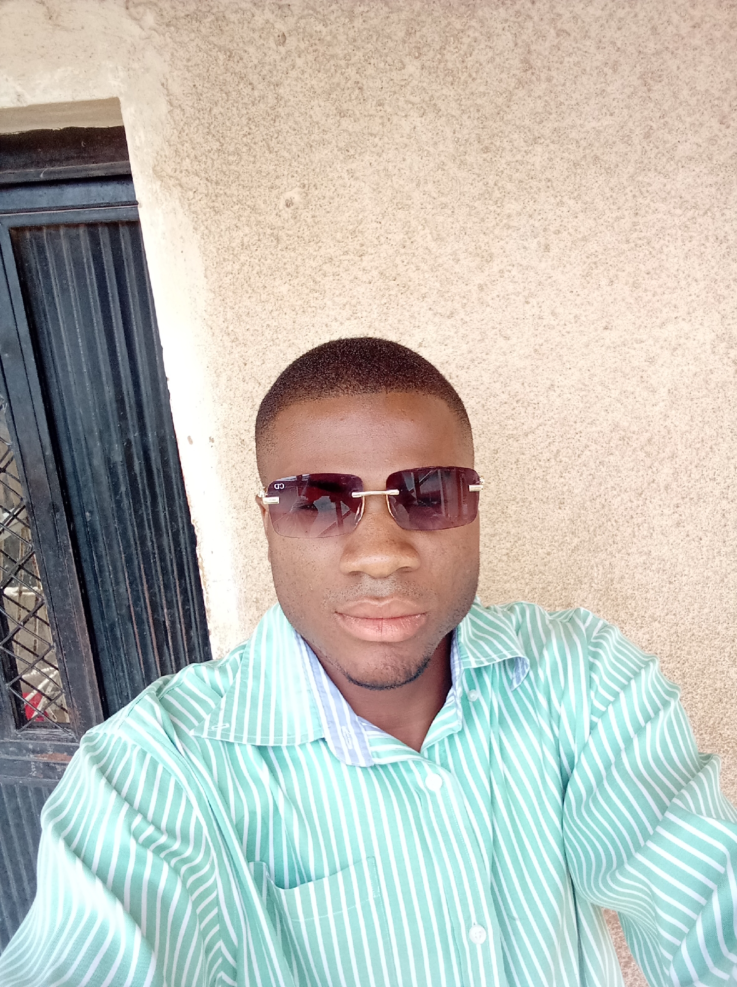 A head-and-shoulders portrait, likely a selfie, features a young man with dark skin, positioned slightly off-center to the right. He has a short, neat haircut and is wearing dark-tinted, rectangular sunglasses with gold-colored frames. A "CD" logo is visible on the left temple of the sunglasses. His facial expression is neutral with his lips slightly parted.
He is dressed in a collared, long-sleeved button-down shirt with vertical light green and white stripes. The shirt appears somewhat casual and slightly wrinkled.
The background consists of two main parts. To the left, behind his left shoulder, is a dark, ribbed metal structure, possibly a security door or window bars, suggesting an exterior or semi-outdoor setting. To the right and extending above his head is a light-colored, textured wall, possibly stucco or concrete with a granular finish, which appears to be a yellowish-beige or off-white. The lighting is bright and even, indicating daytime conditions, likely sunny, which is consistent with the sunglasses. The image evokes a casual and relaxed mood in what is contextually described as Jos, Nigeria.