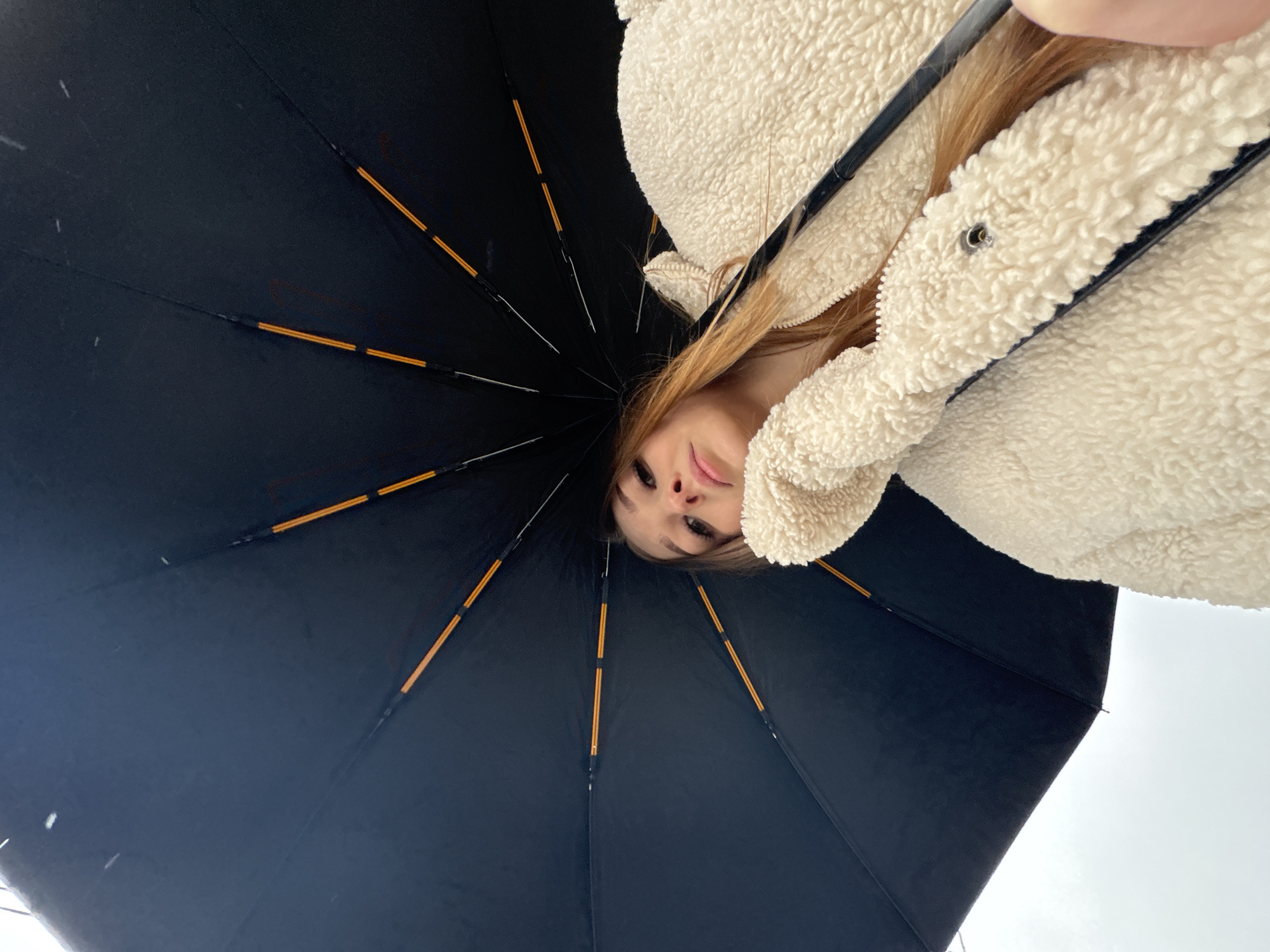 A low-angle, close-up shot depicts a person holding a black umbrella in what appears to be a snowy or wet environment. The person, presumably female, is partially visible, with fair skin, light brown hair, and wearing a light-colored, fleece jacket. The umbrella's internal frame features black ribs with orange accents. Snowflakes or raindrops are visible against the overcast sky. The location cannot be ascertained from the image.