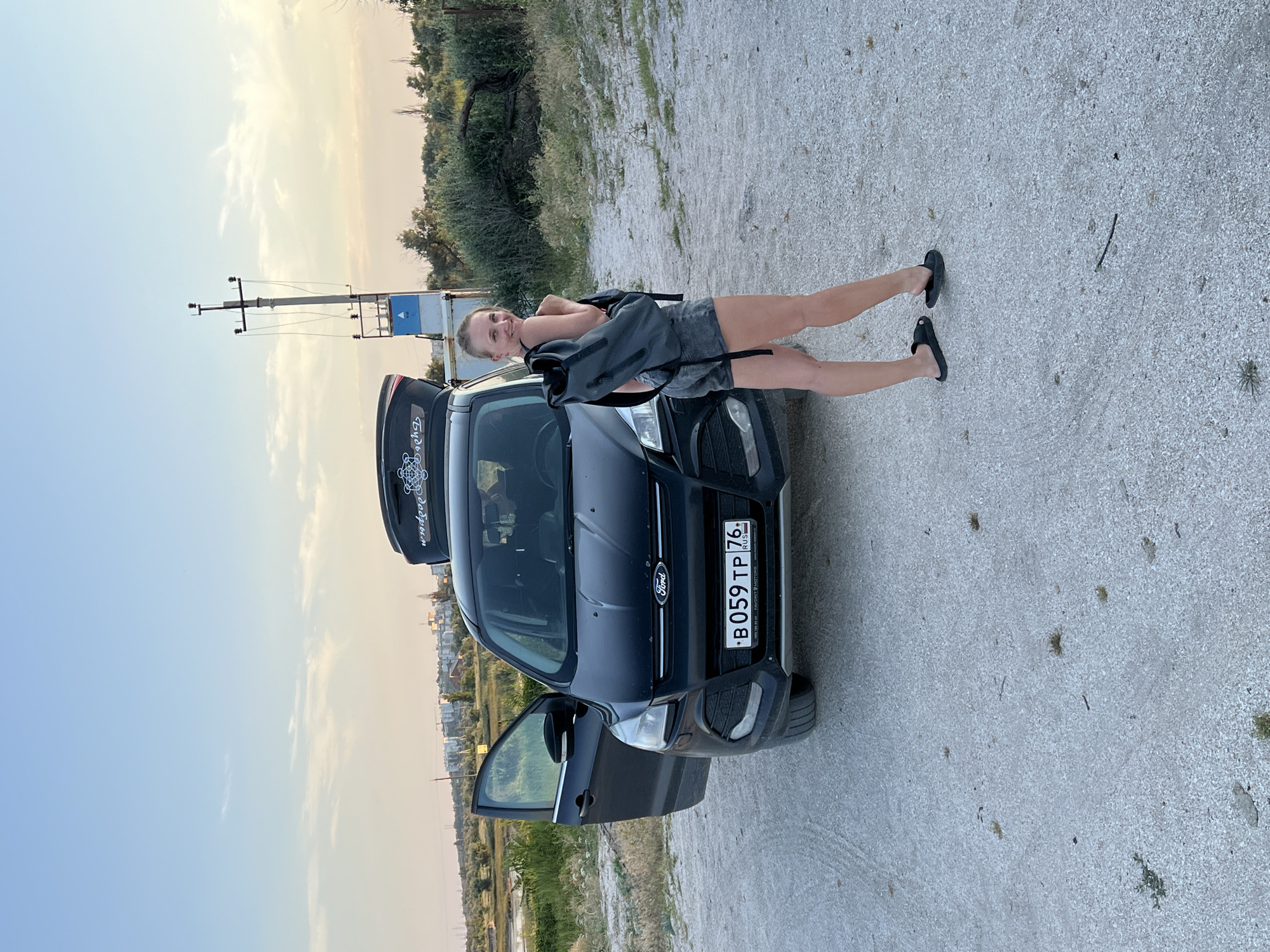 A woman with blonde hair pulled back is posing next to a dark blue Ford SUV. She is wearing a gray tank top and denim shorts, and has a gray backpack slung over her shoulders. Her legs are extended and she is standing with her feet flat on the ground. The car's rear hatch is open, revealing the interior. The license plate on the front of the car reads "B059 TP 76 RUS". The scene is outdoors, with a gravelly ground and some sparse vegetation in the background. A utility pole with electrical equipment is visible to the left of the car. The sky is a pale blue with wispy clouds, suggesting it is late afternoon or early evening. The overall mood appears relaxed and casual. The location is identified as Мысовское сельское поселение, Ukraine.