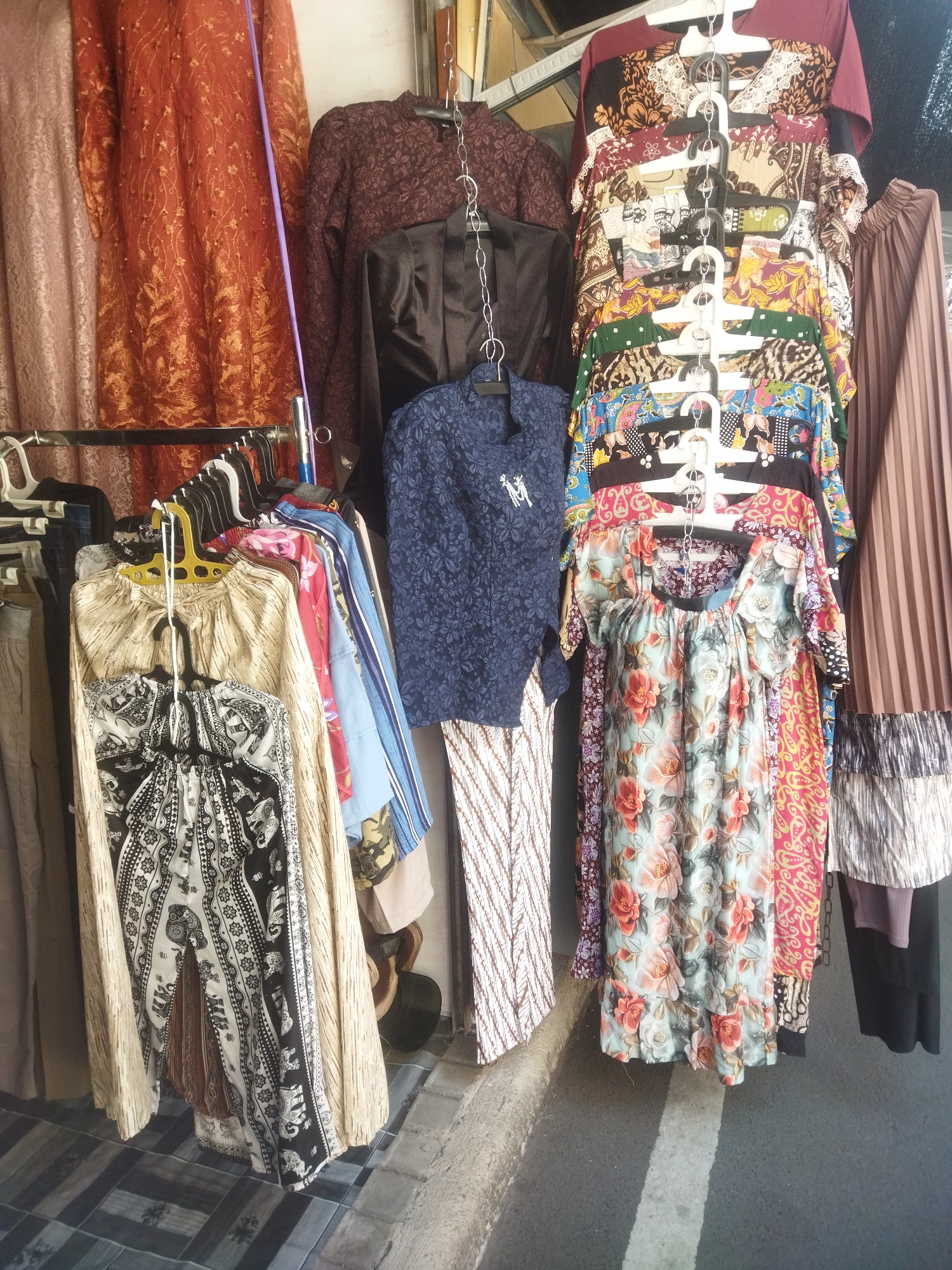 This image captures a vibrant display of women's clothing hanging outside a store in the city of Wonogiri, Indonesia. The clothing includes various styles and patterns, such as floral prints, batik designs, and solid colors. The garments are neatly arranged on hangers and racks, creating an eye-catching presentation. The scene appears to be a bustling street market or a small boutique. The background features a building facade with a white wall and a gray sidewalk, typical of Indonesian urban settings. The lighting suggests it is daytime, with shadows indicating sunlight. The overall impression is one of a lively and colorful marketplace, offering a diverse selection of traditional and contemporary Indonesian fashion.
